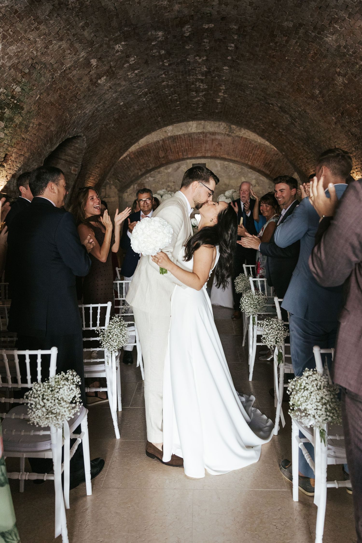 Newlyweds share their first kiss during intimate ceremony in rustic brick-vaulted venue with guests looking on.