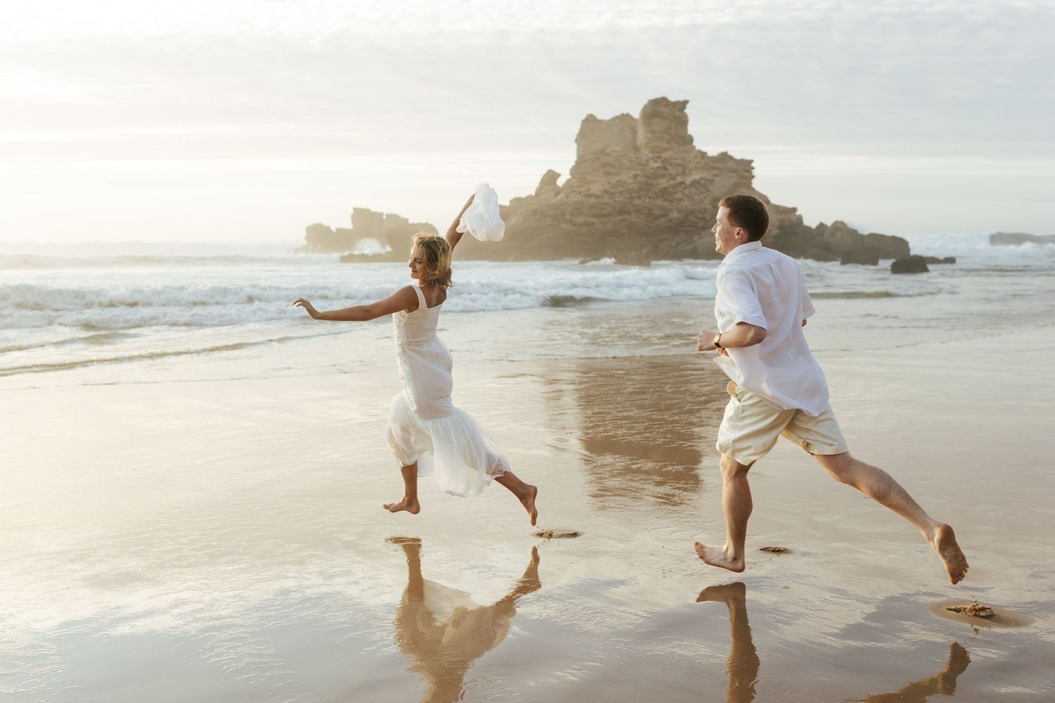 Couple runs playfully along a sandy beach at sunset with a rocky formation in the background. Praia do Castelejo, Portugal.