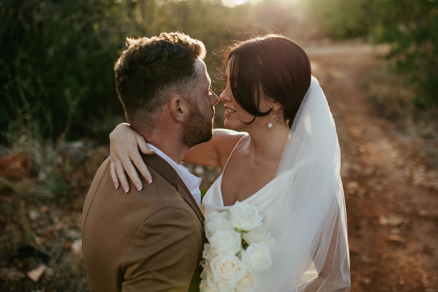 Newlywed couple embraces on a dirt path at golden hour as bride holds white bouquet in romantic sunset moment.