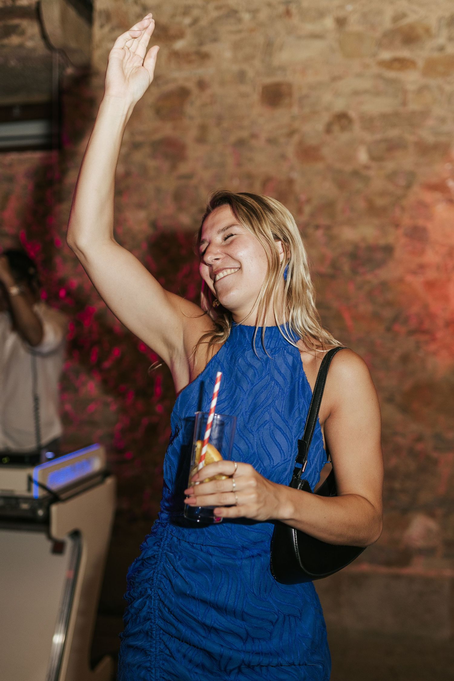 A person in a blue sleeveless dress dances joyfully with one arm raised while holding a drink at a party.