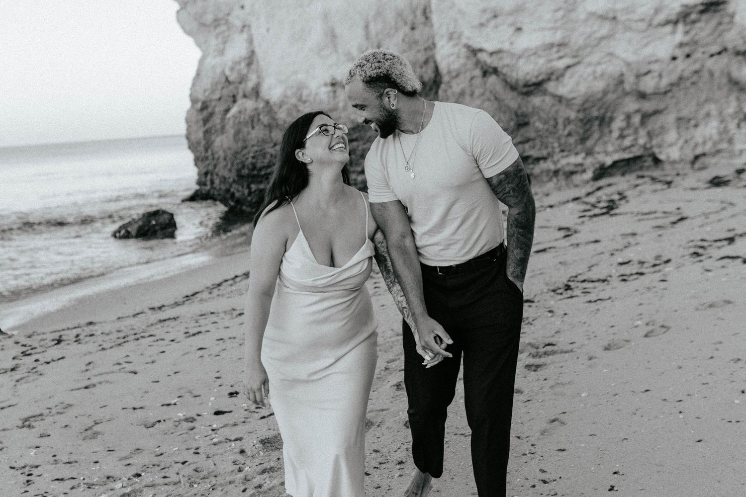 A romantic black and white beach photograph of a couple by cliffside rocks holding hands on the shoreline.
