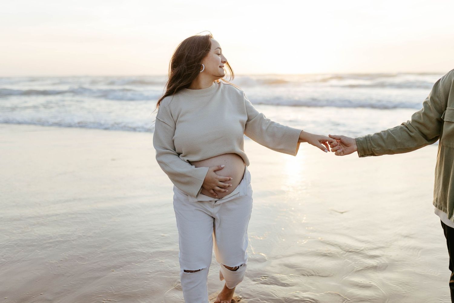 A couple holds hands while walking along a beach at sunset with gentle waves in the background.