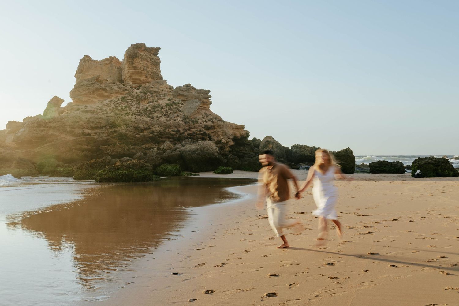 Two people in white clothing run together along a scenic beach with dramatic rock formations and cliffs during golden hour.
