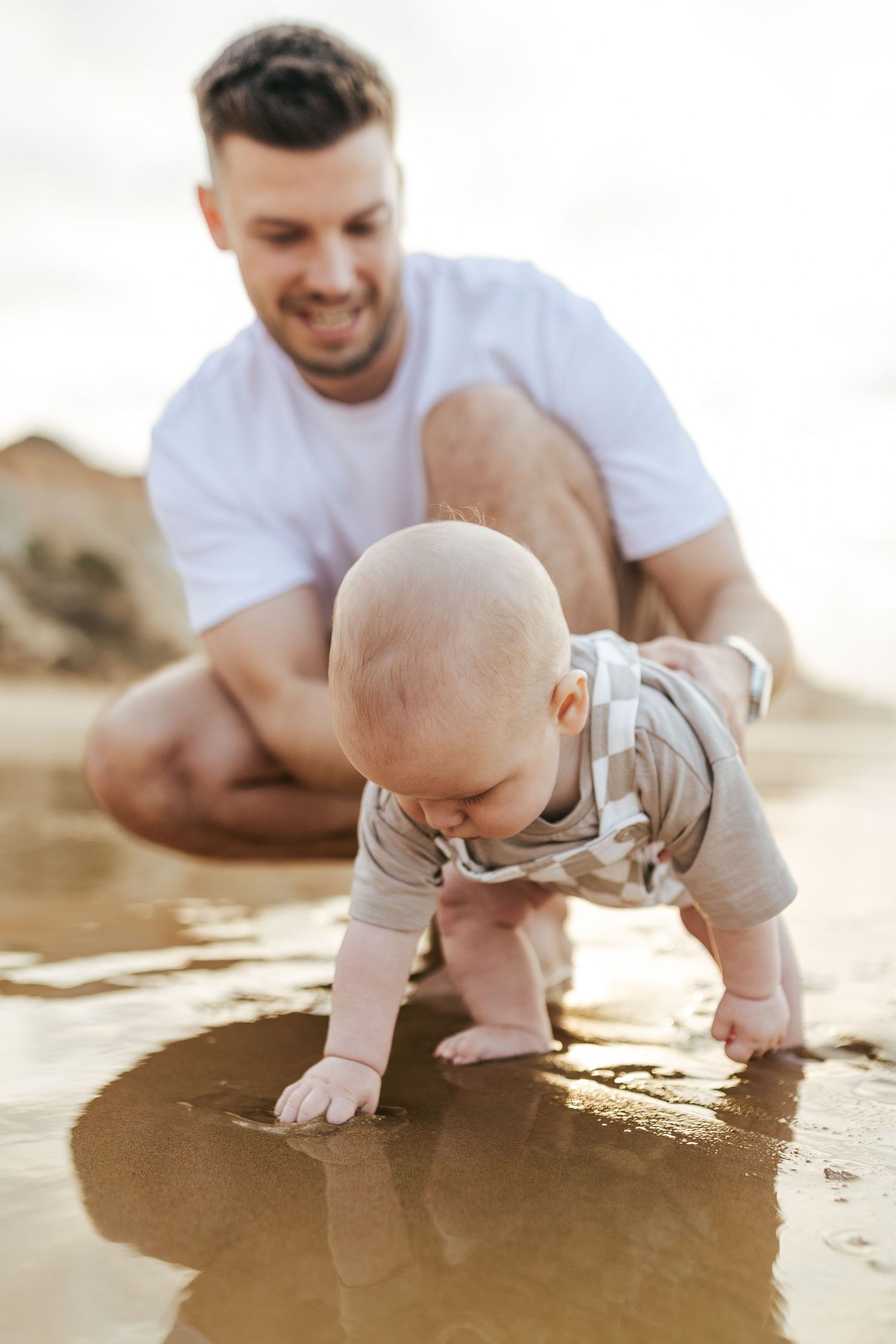 A baby crawls on wet sand at the beach while an adult watches during golden hour sunset light.