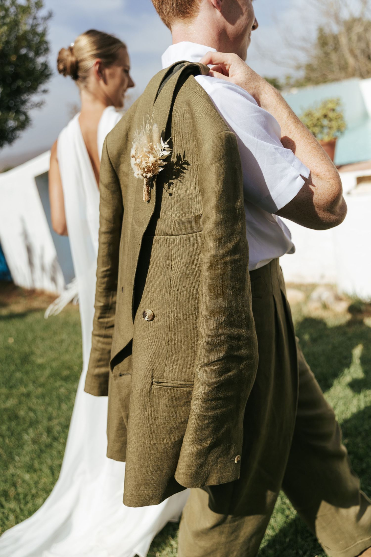 Groom in olive linen suit carries jacket with floral boutonniere while bride in backless white gown stands nearby outdoors.
