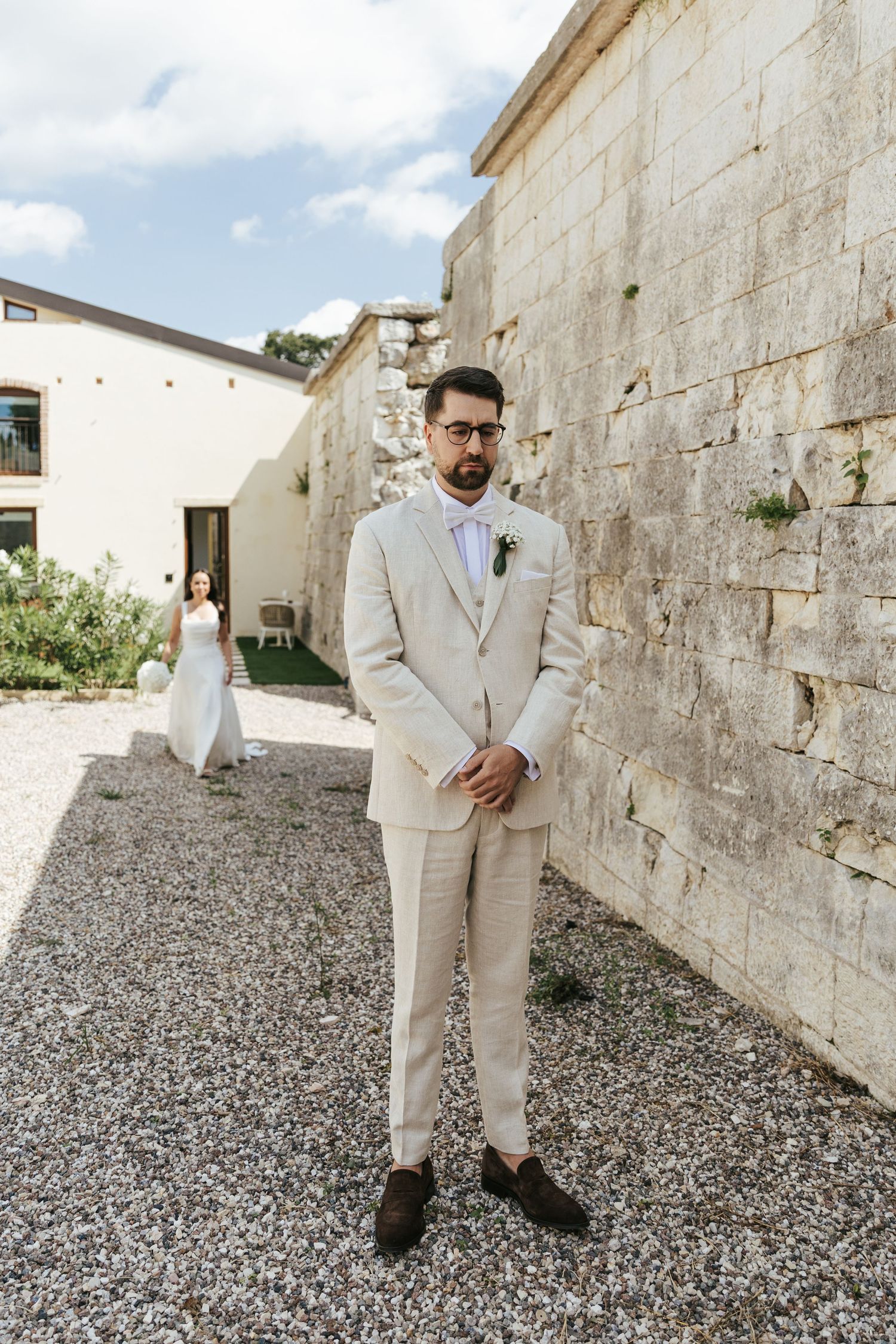 A groom in a light beige suit poses against stone walls on a gravel path.