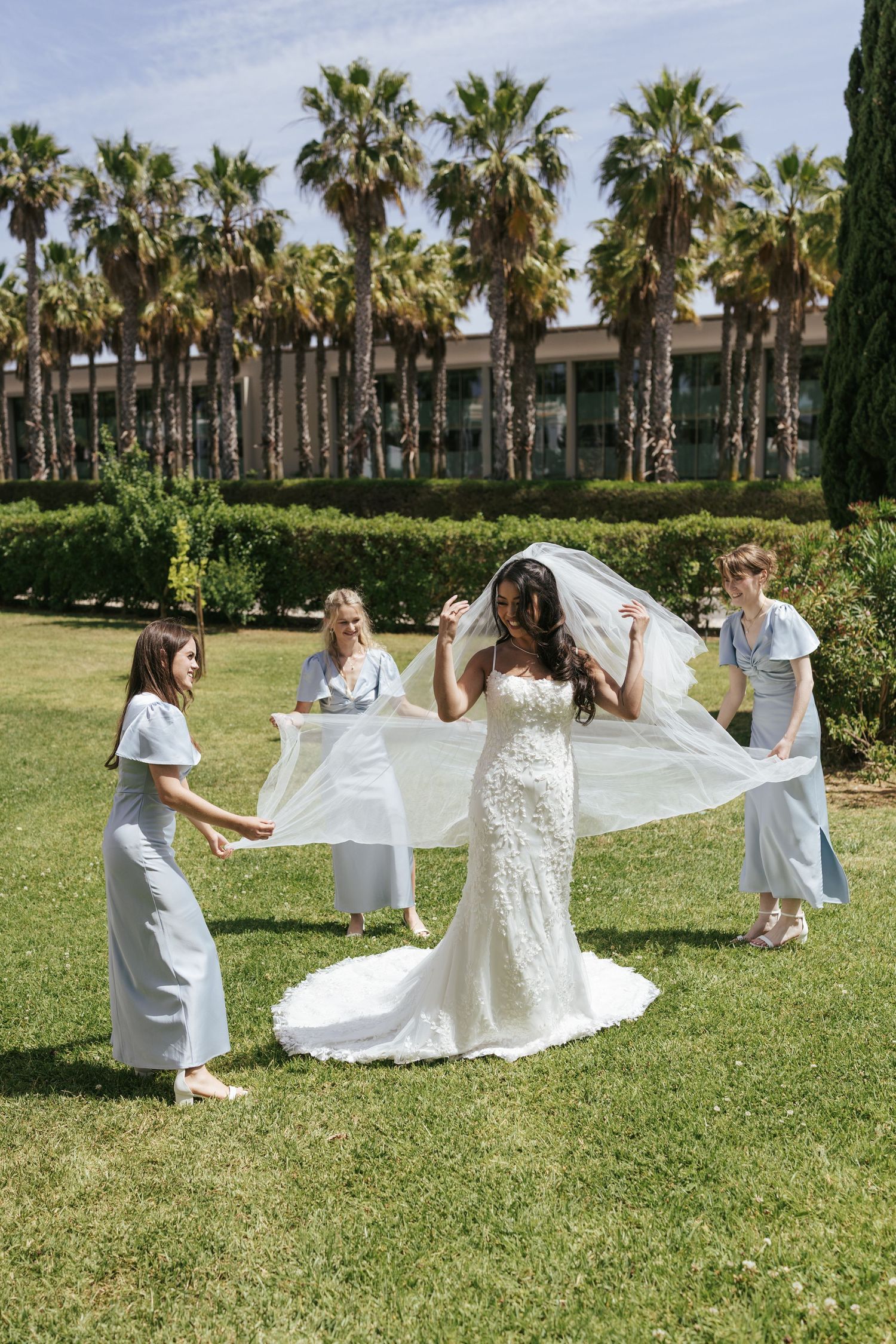 Bride in white lace gown with bridesmaids in blue dresses holding veil on sunny garden lawn with palm trees.