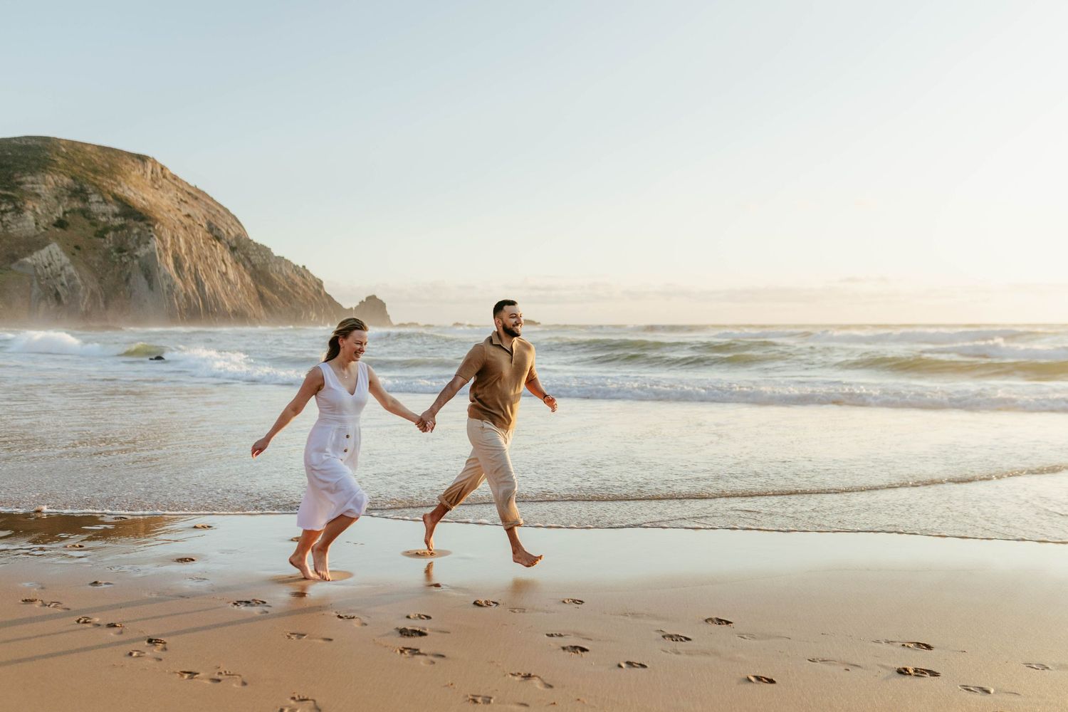Two people holding hands while running along a scenic beach at sunset with cliffs in the background. Praia do Castelejo, Sagres, Portugal.
