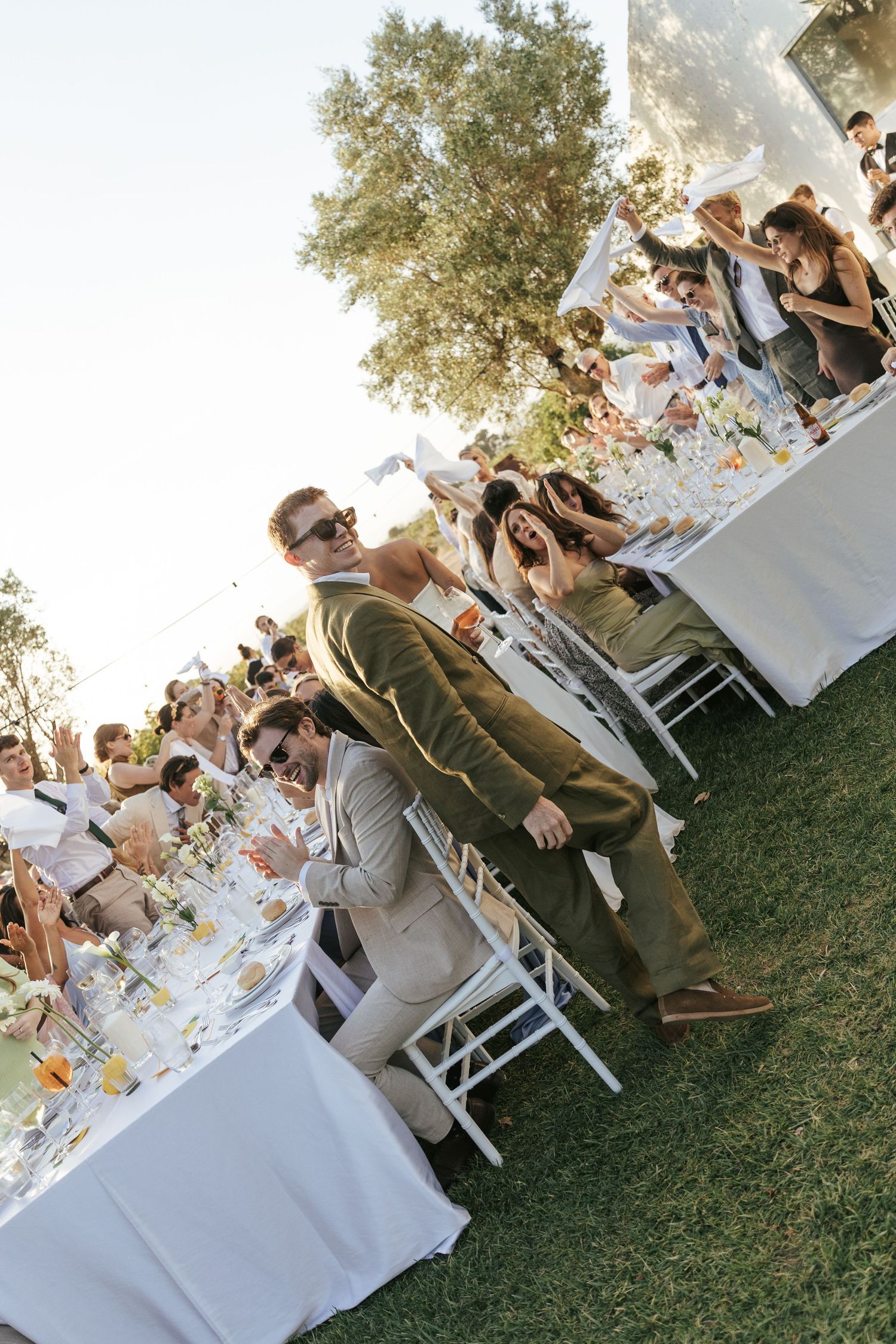 Guests seated at elegant outdoor dining tables with white linens under a large tree during a daytime celebration.