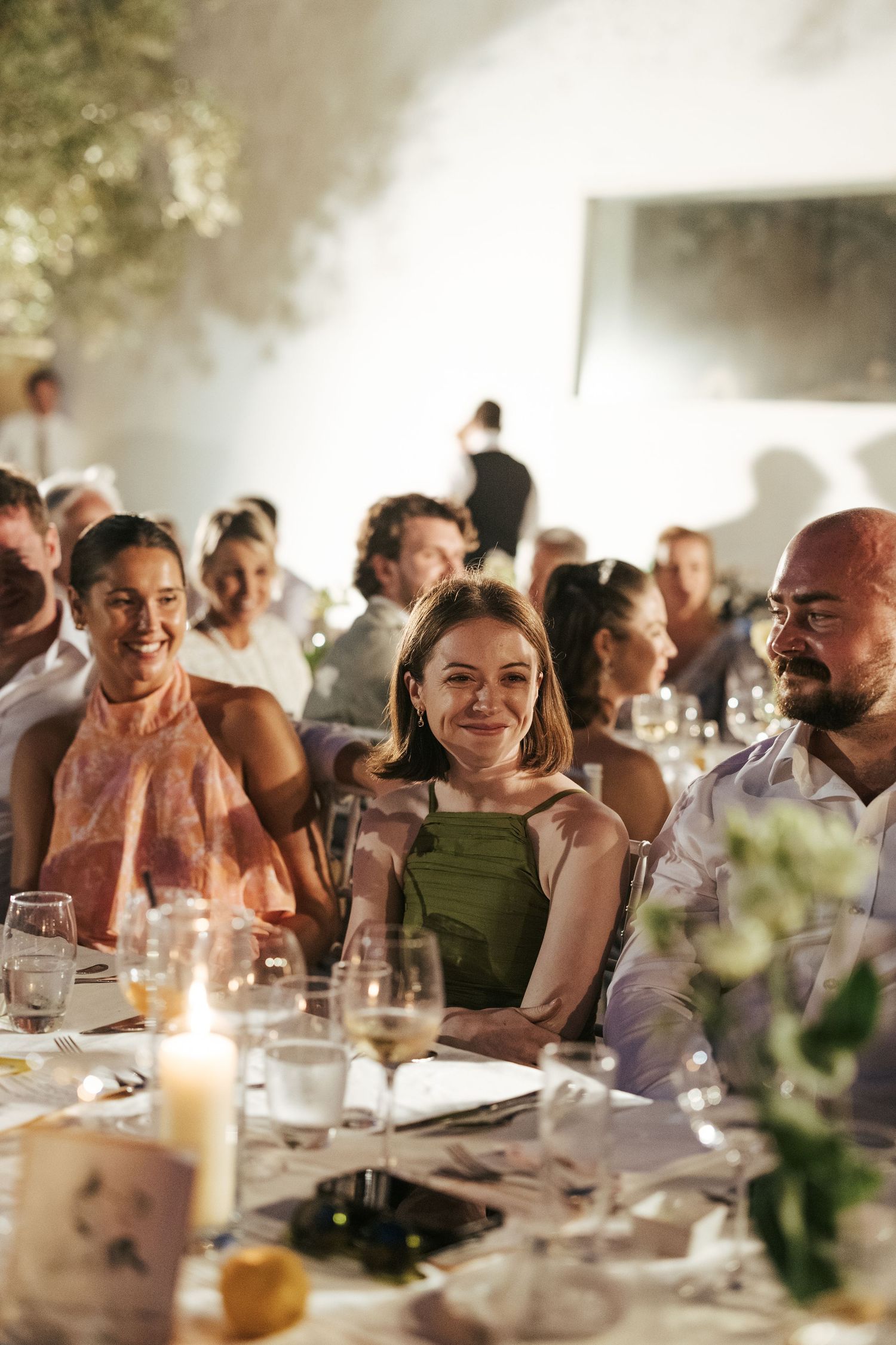 Smiling wedding guests seated at elegantly decorated reception dinner table with candles and glassware.