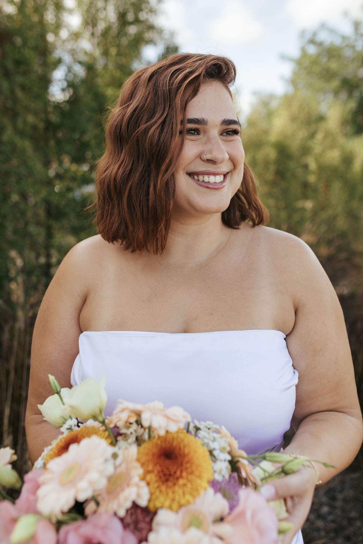 A person in a white strapless dress holds a vibrant bouquet of summer flowers outdoors.