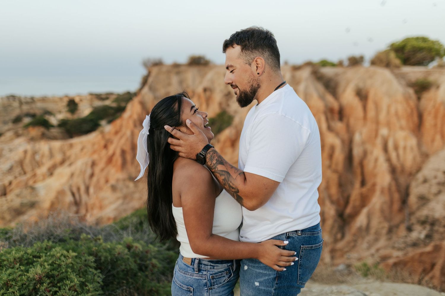 A romantic couple embraces against a backdrop of rugged desert cliffs and greenery at sunset.