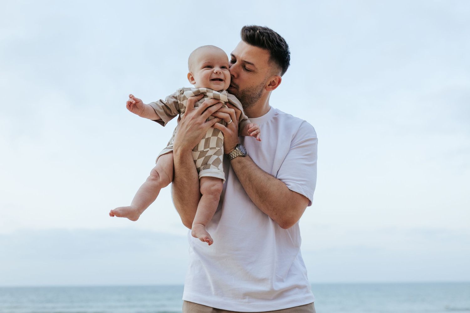 A tender moment of affection between parent and baby outdoors against an ocean backdrop.