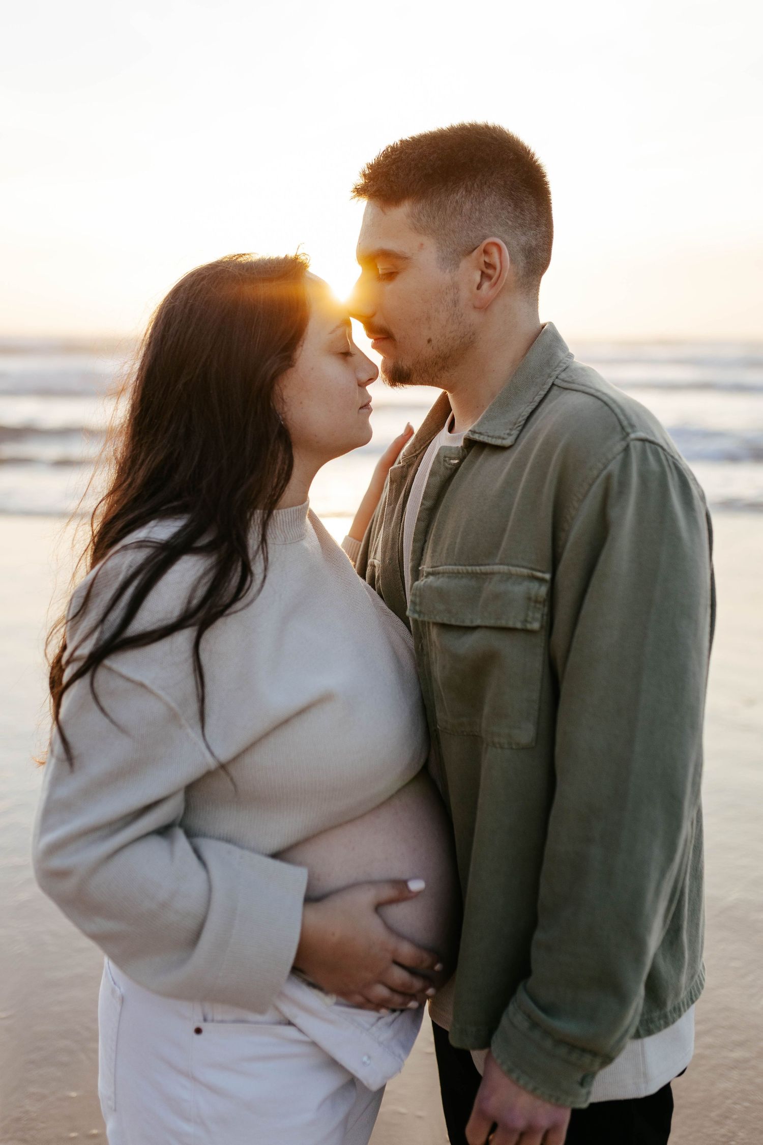 A romantic couple shares an intimate moment on the beach at sunset with sunlight glowing between them.