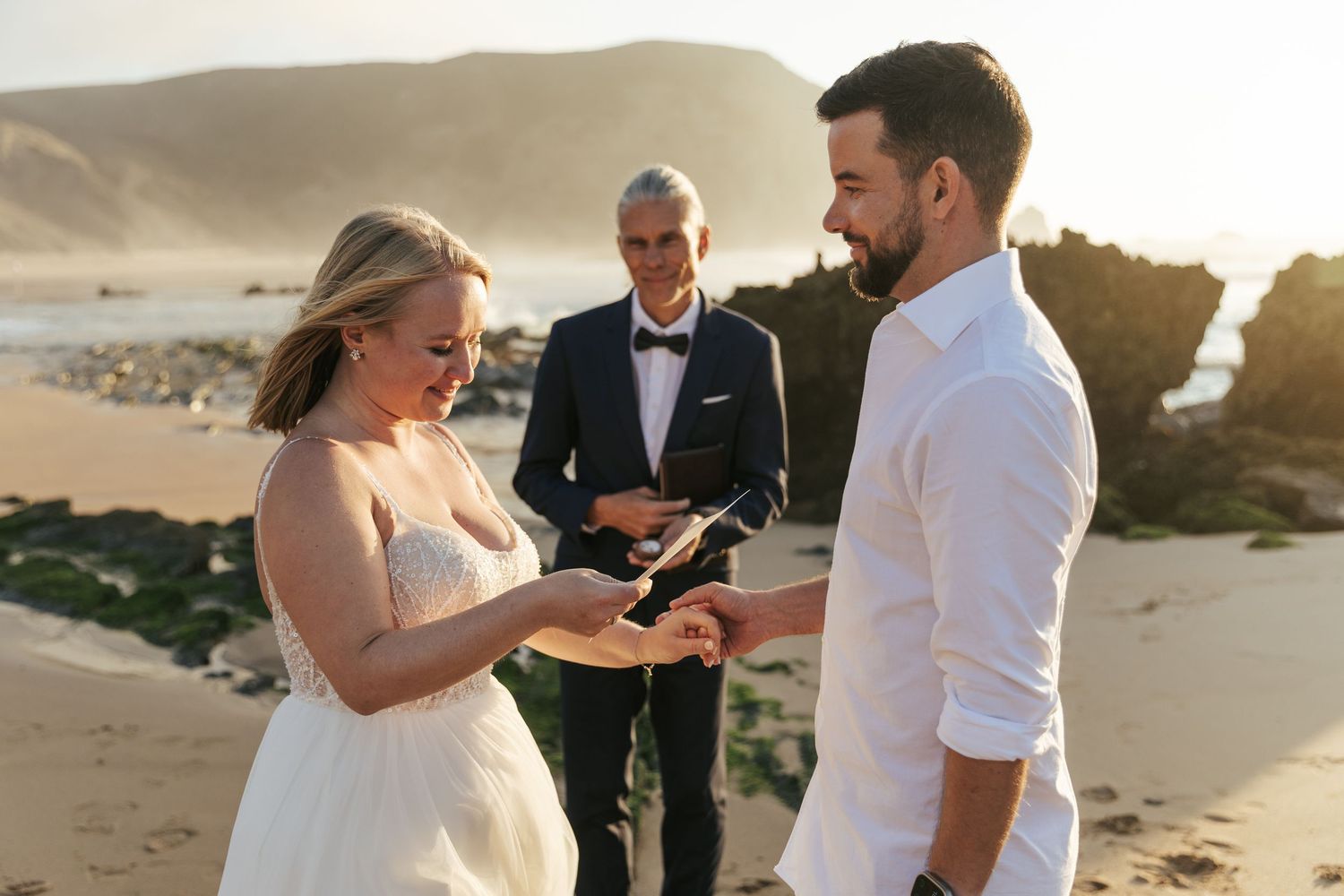 A couple exchanges vows during an intimate beach wedding ceremony at sunset with rocky cliffs in the background.