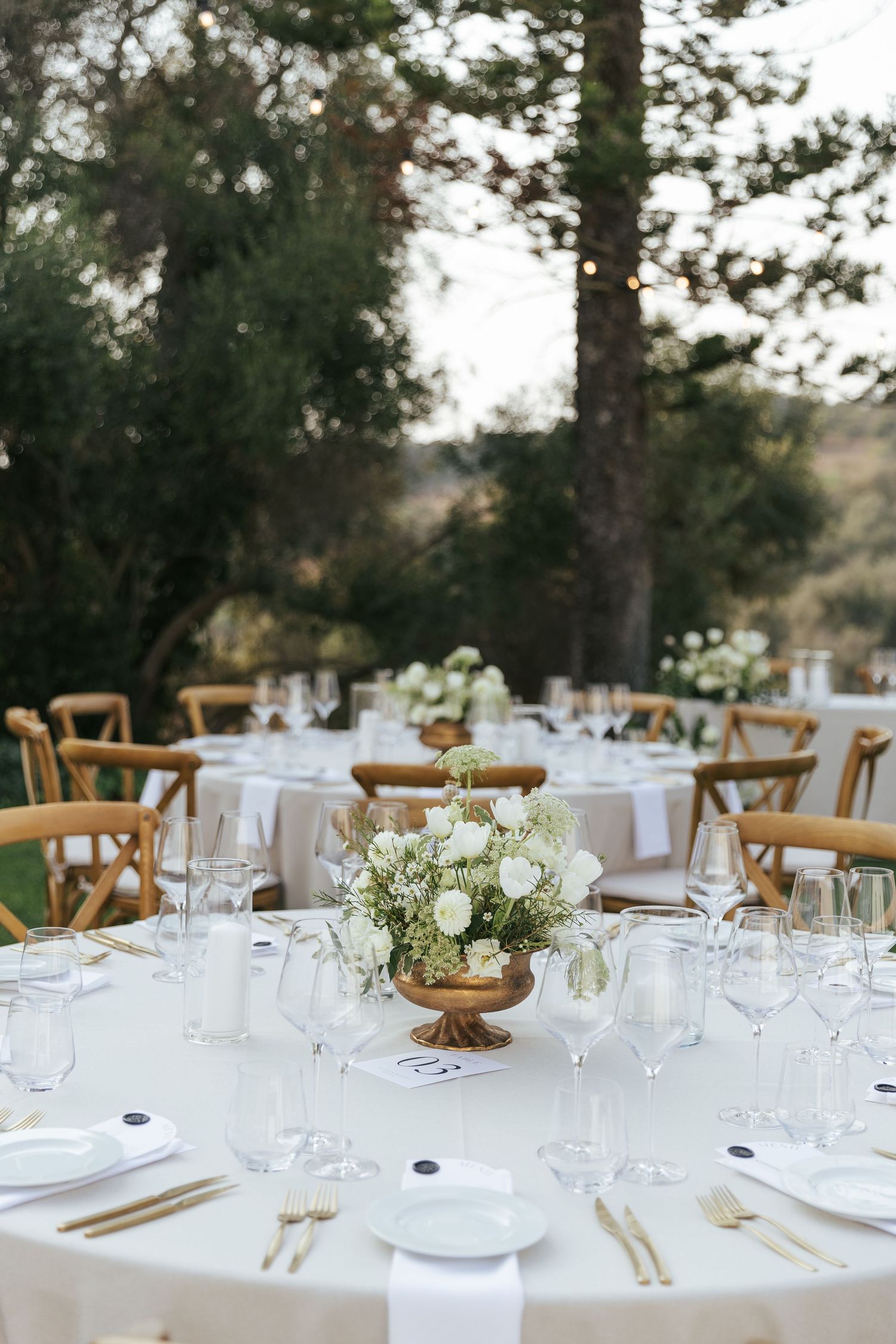 Elegant outdoor wedding reception table setting with white linens and delicate floral centerpieces.