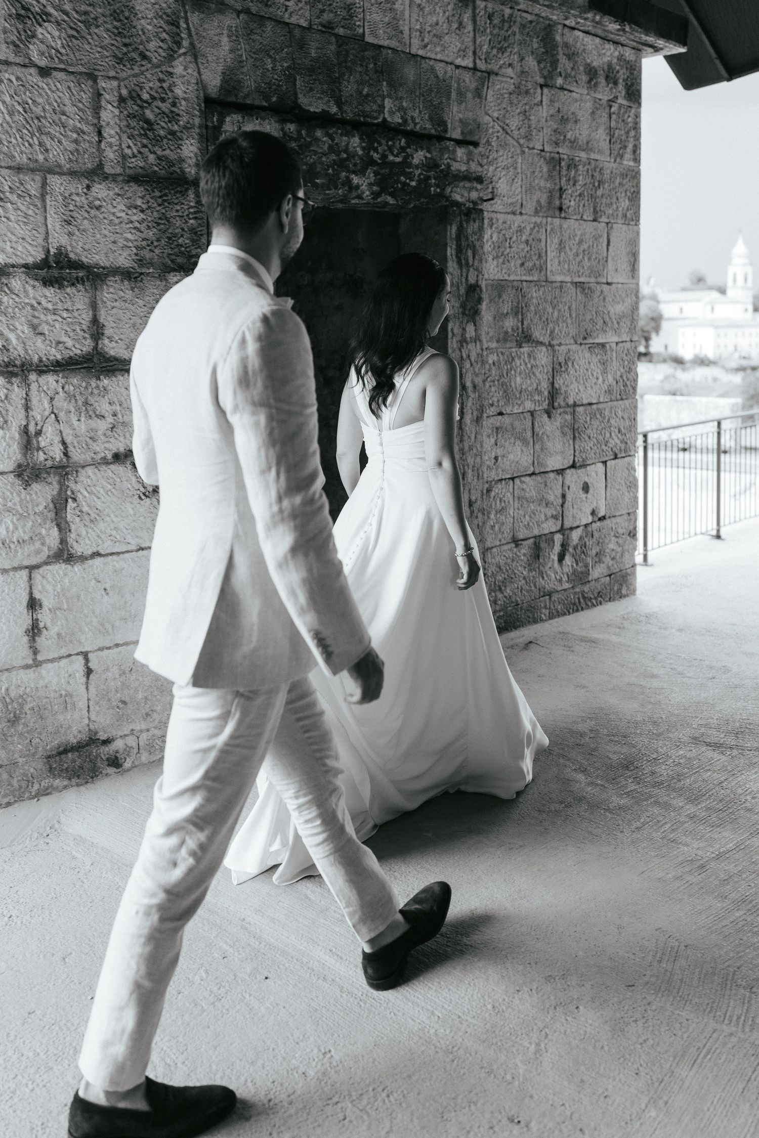 Couple in white attire walking through stone archway during wedding photoshoot.