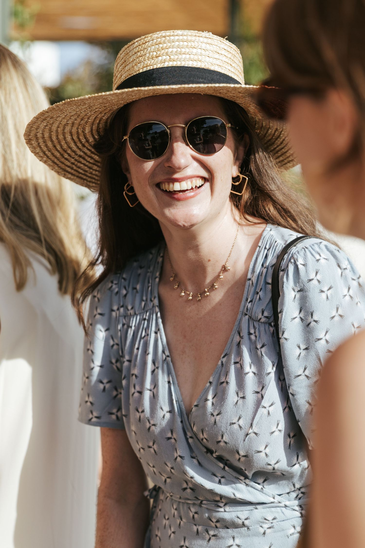 Smiling woman in straw hat and sunglasses wearing blue floral dress at outdoor summer event.