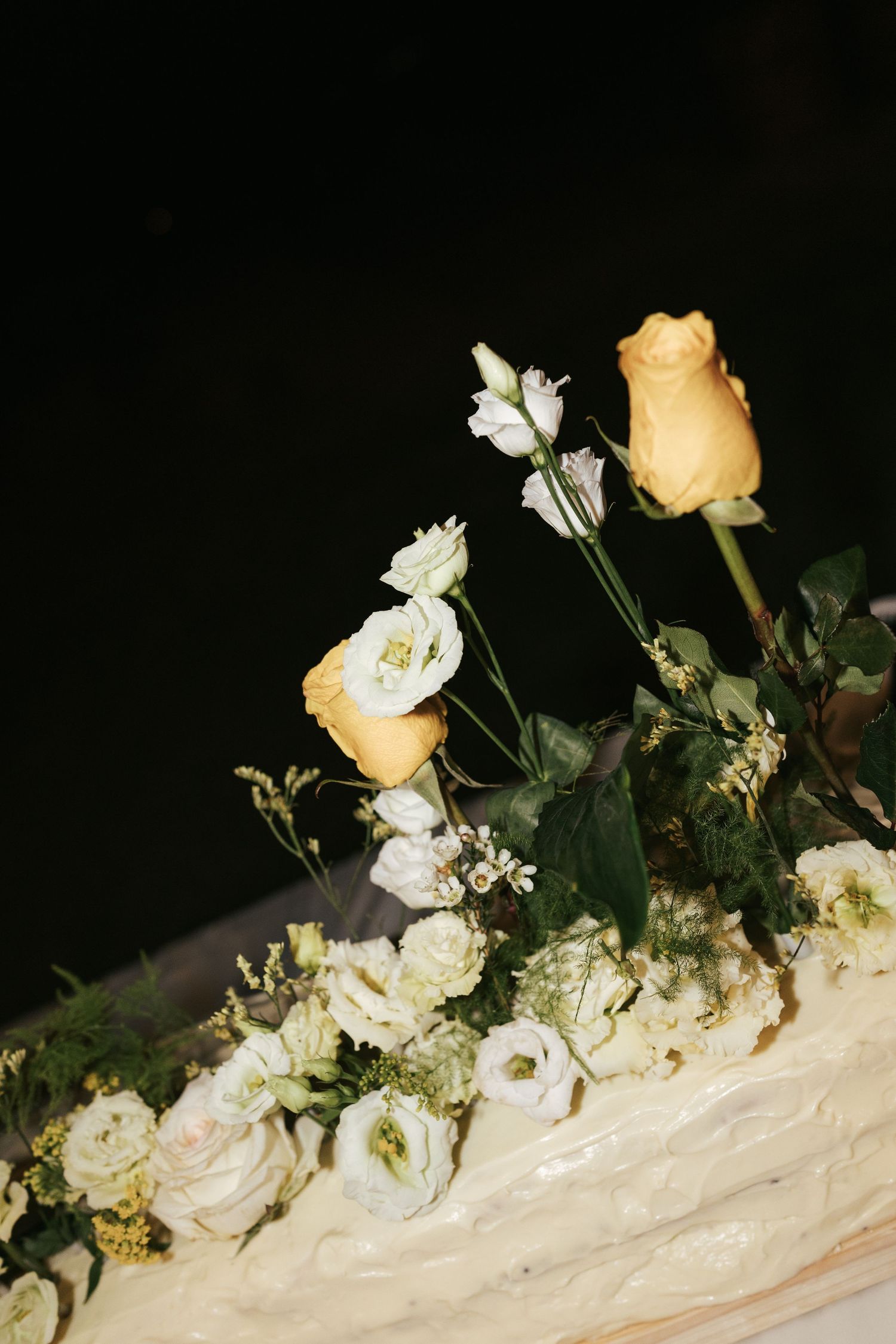 Elegant wedding cake decorated with yellow roses and white flowers on dark background.