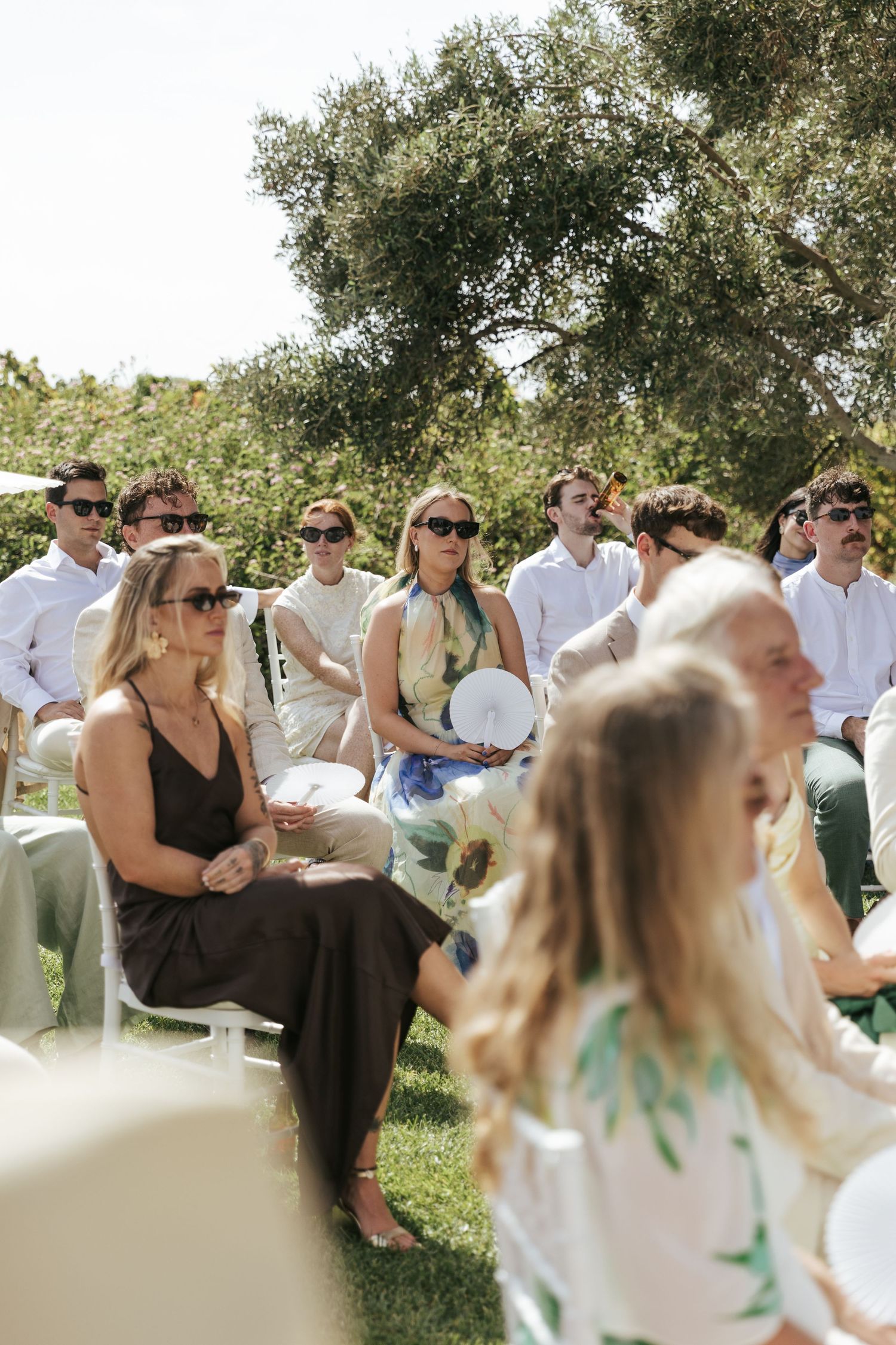 Guests seated outdoors at a sunny wedding ceremony, wearing sunglasses and elegant attire on white chairs.