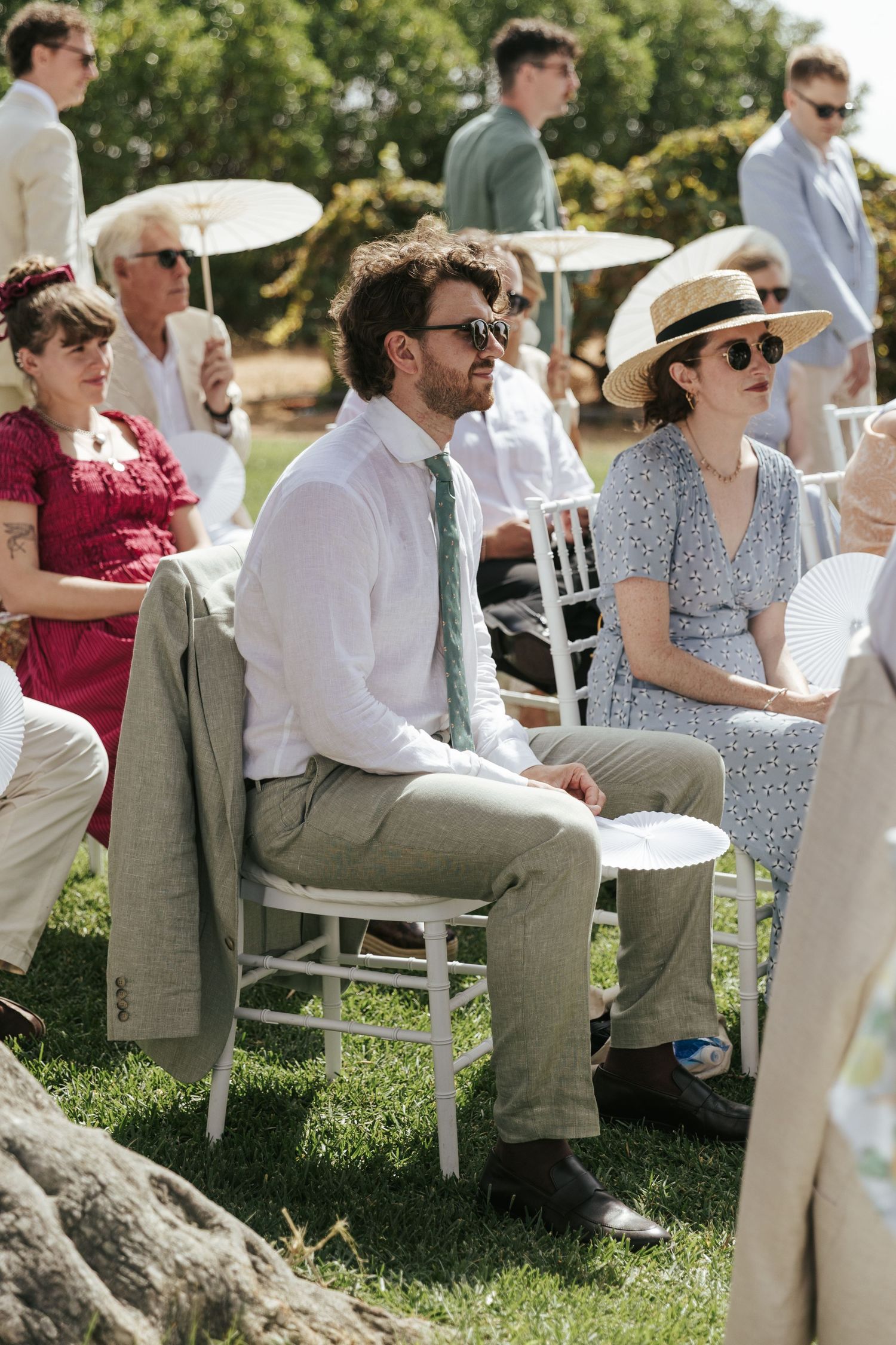 Wedding guests seated outdoors in summer attire, including sunglasses and straw hat, watching ceremony.