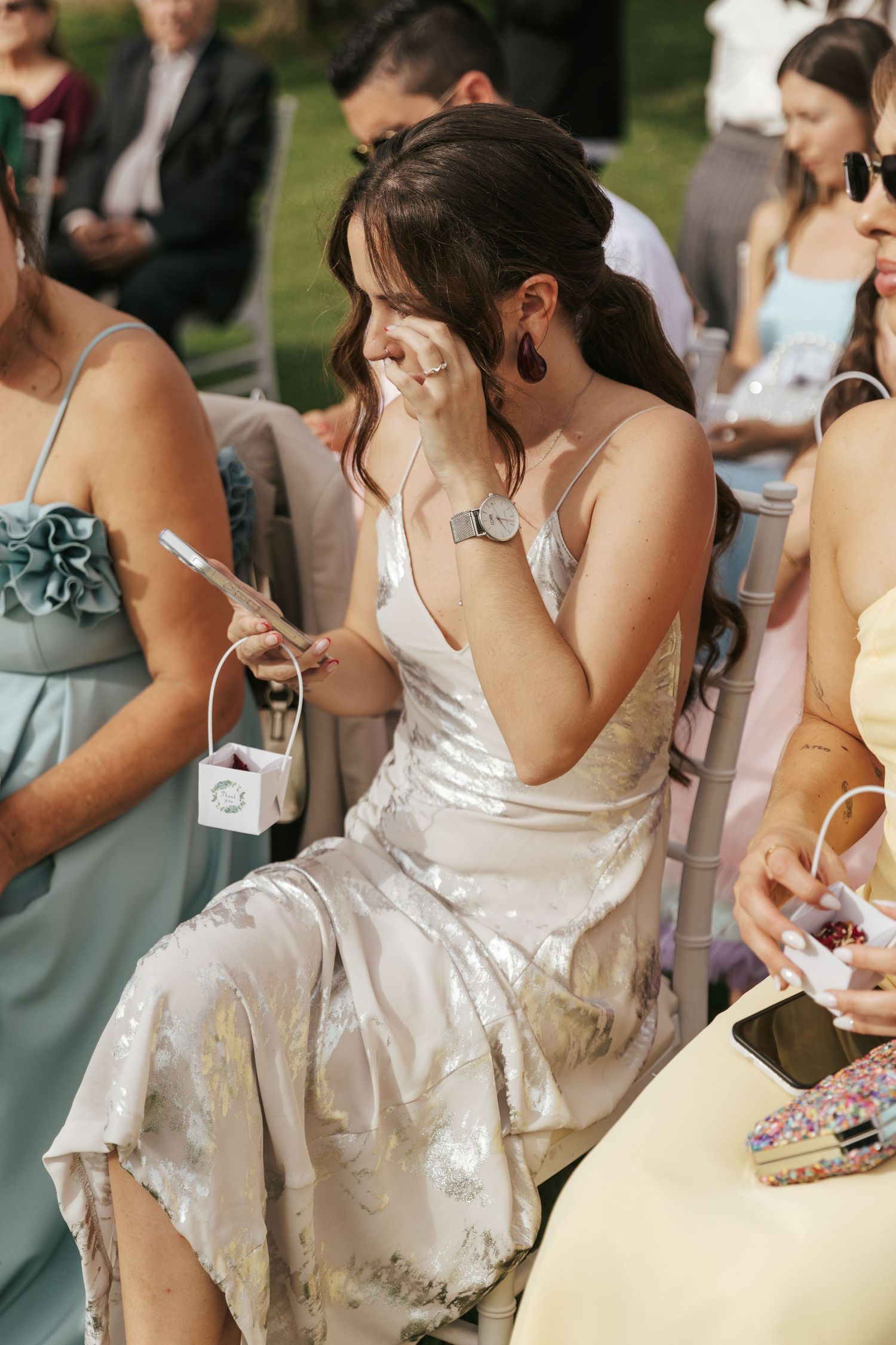 Wedding guest in silver satin dress wipes away tears during an emotional moment at outdoor ceremony.