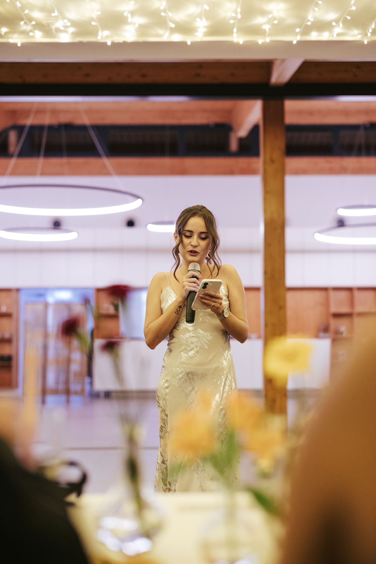 Someone in a champagne-colored dress gives a speech with a microphone at an elegant indoor reception under string lights.
