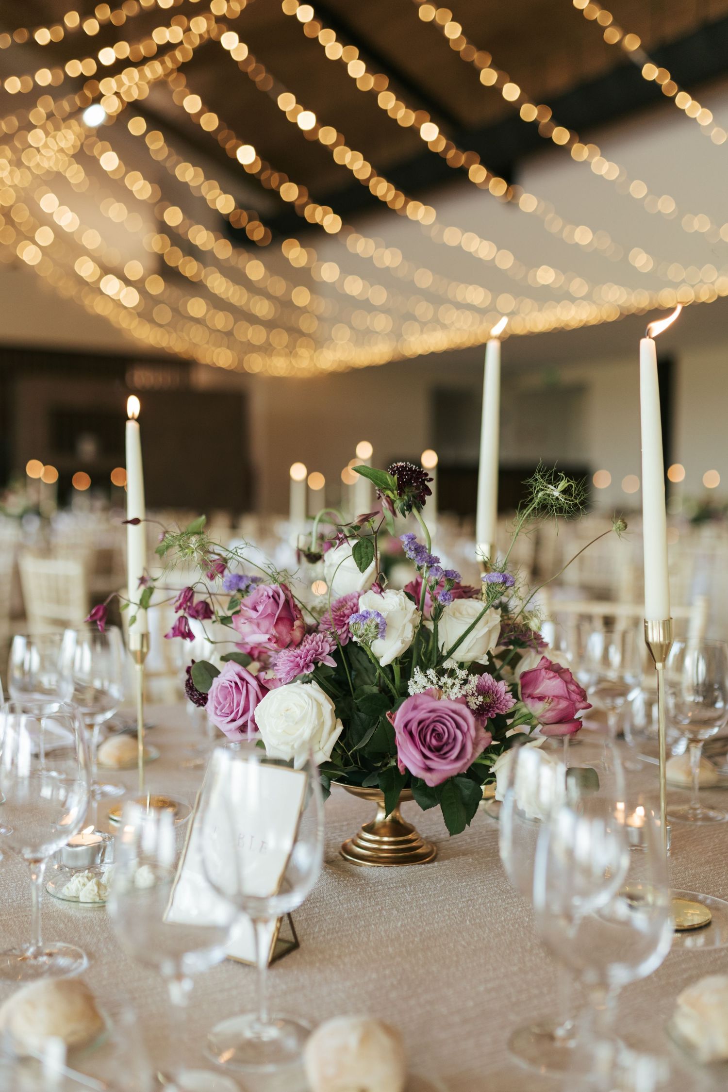 Elegant table setting with purple and white rose centerpiece under romantic string lights at a wedding reception.