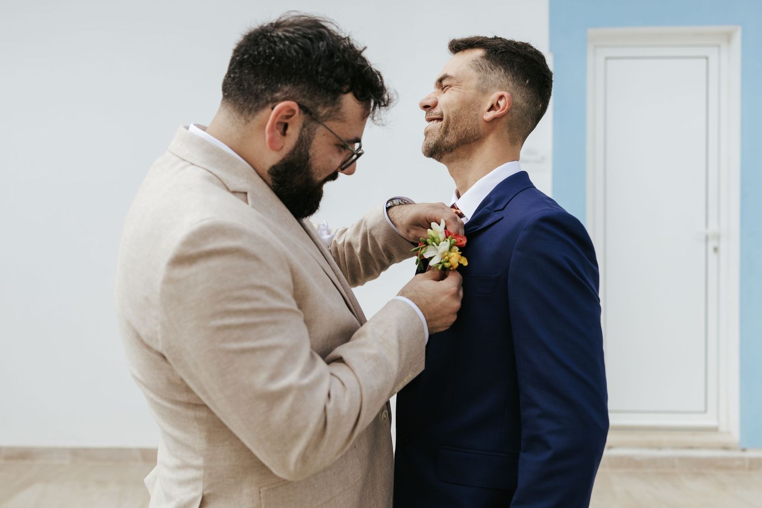 Two people in formal attire, one in beige and one in navy, share a tender moment while adjusting a boutonniere.