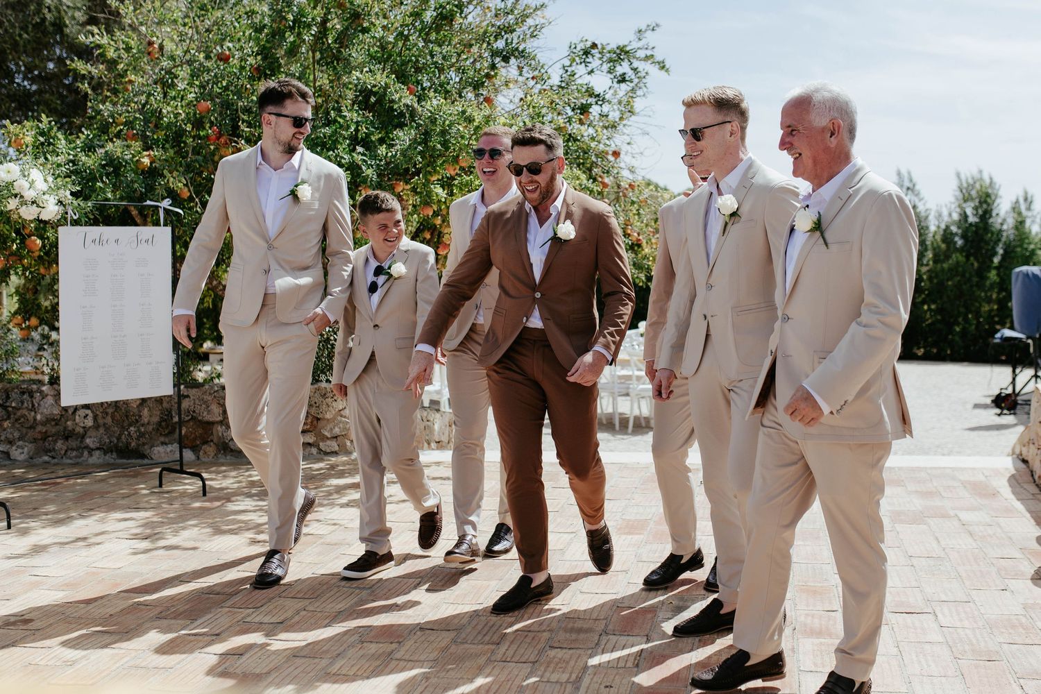 Groomsmen walking together outdoors in coordinated suits of beige and brown tones during a sunny wedding day.