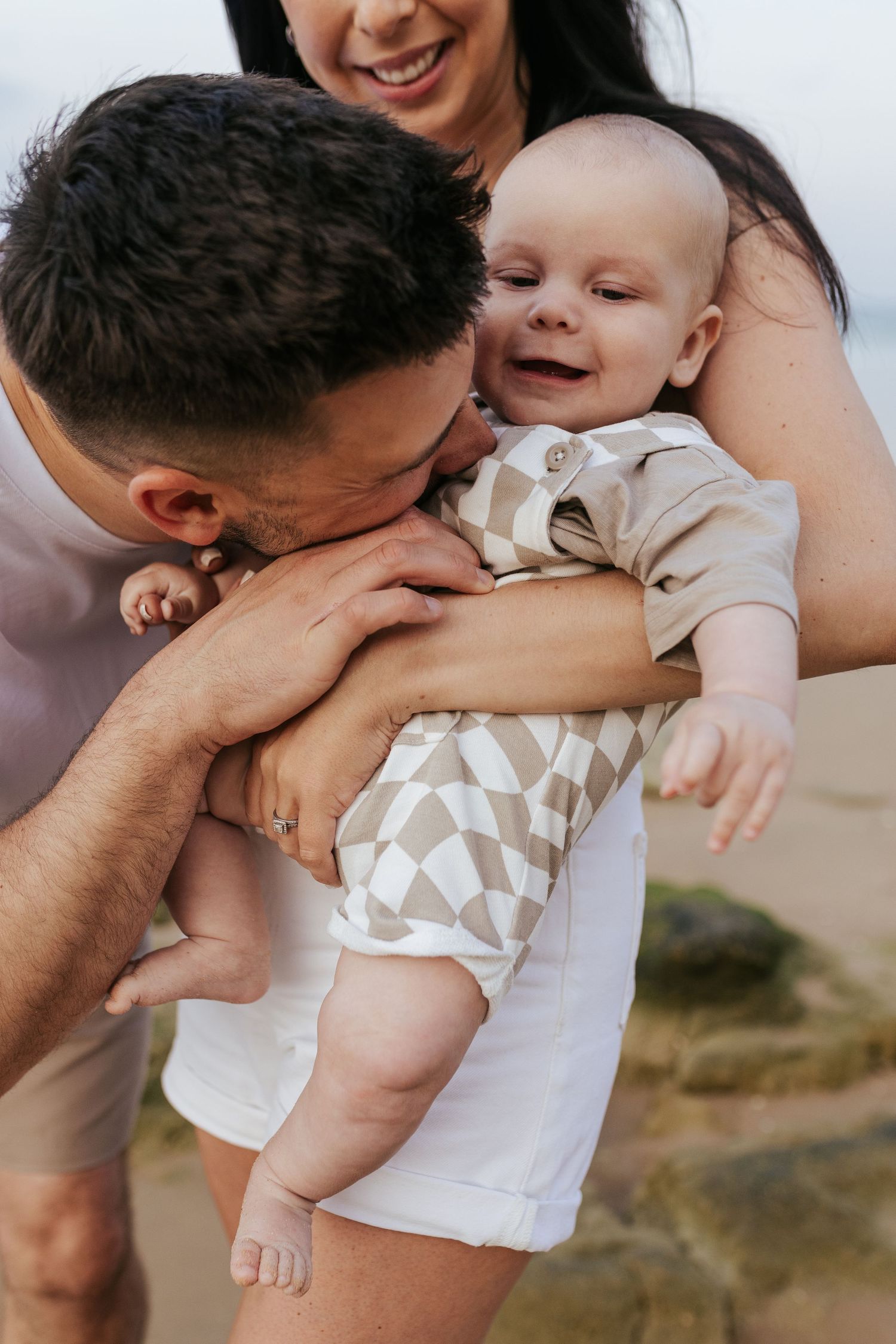 A happy baby laughs while being held close by parents in a warm outdoor setting during a tender family moment.
