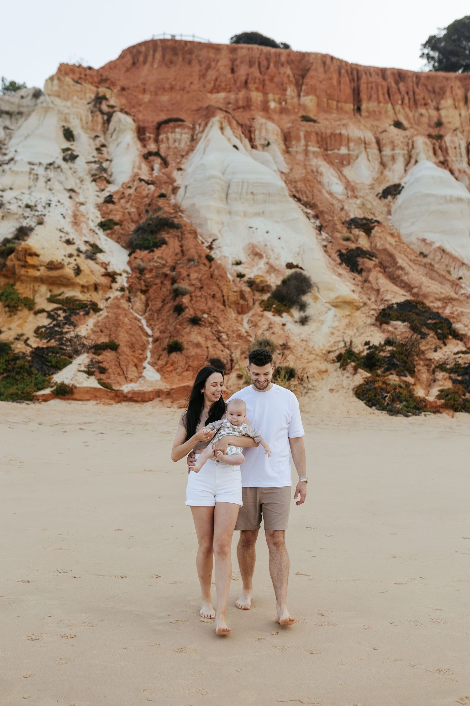 A couple walks barefoot along a beach with dramatic red and white cliffs rising behind them on a sunny day.