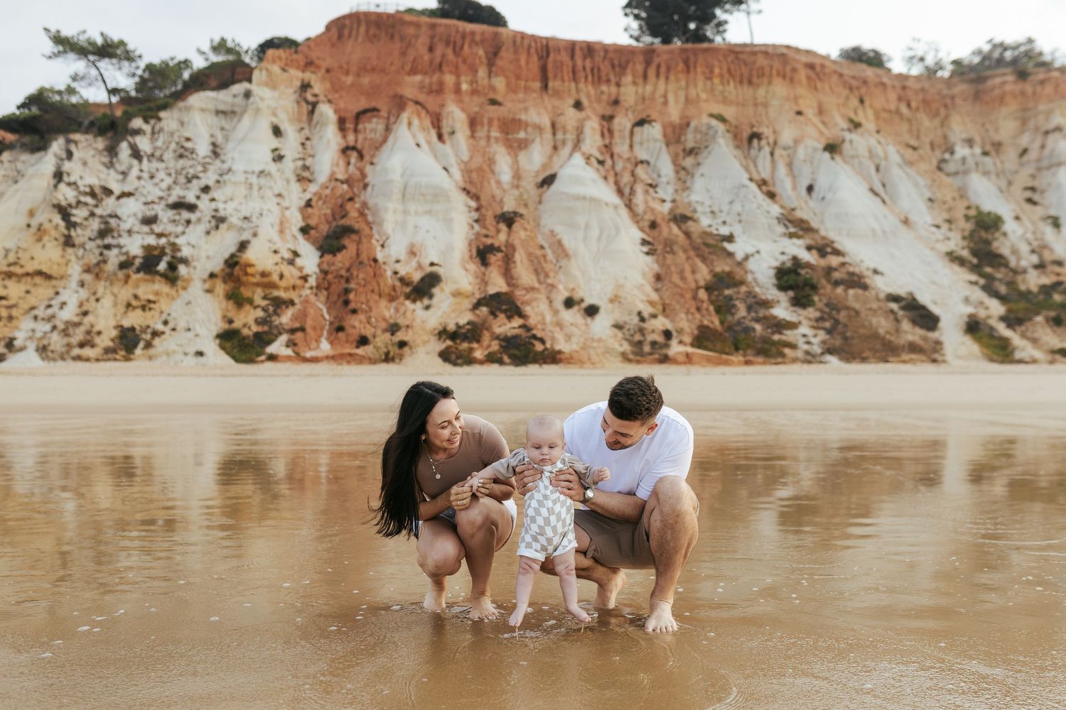 Two people playing with a dog by the water near colorful clay cliffs at a beach.
