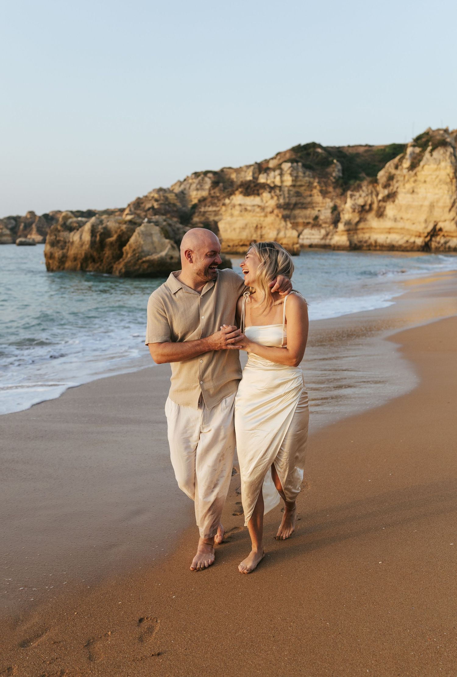 A couple walks barefoot along a sandy beach with dramatic rocky cliffs and ocean waves in the background at sunset.