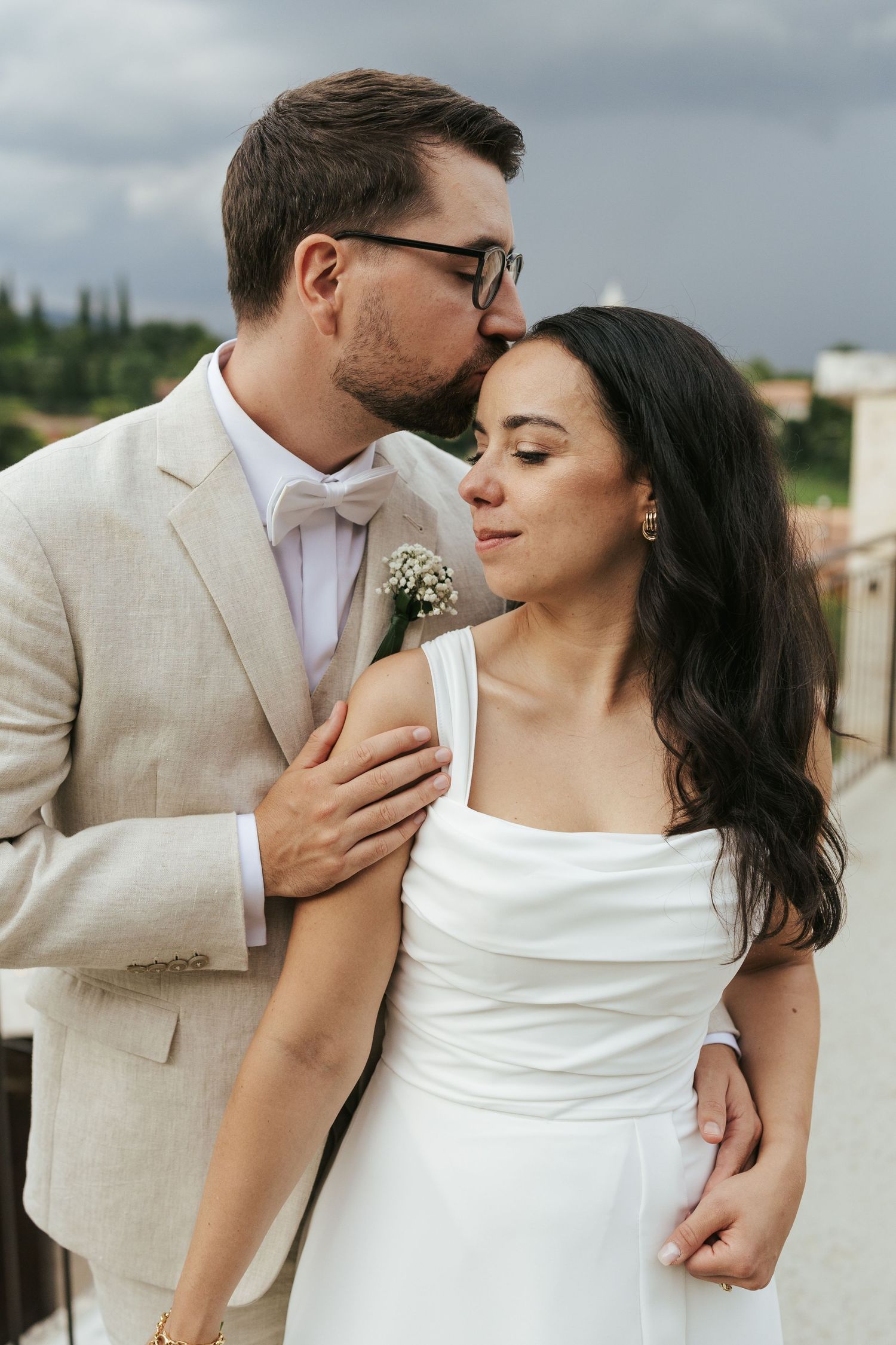 Intimate moment between newlyweds on terrace with cloudy sky and scenic backdrop.