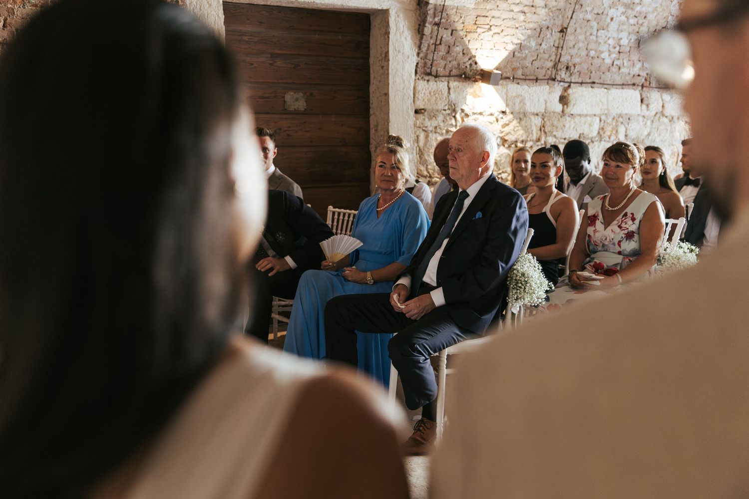 Wedding ceremony guests seated in rows inside historic brick venue with natural lighting.