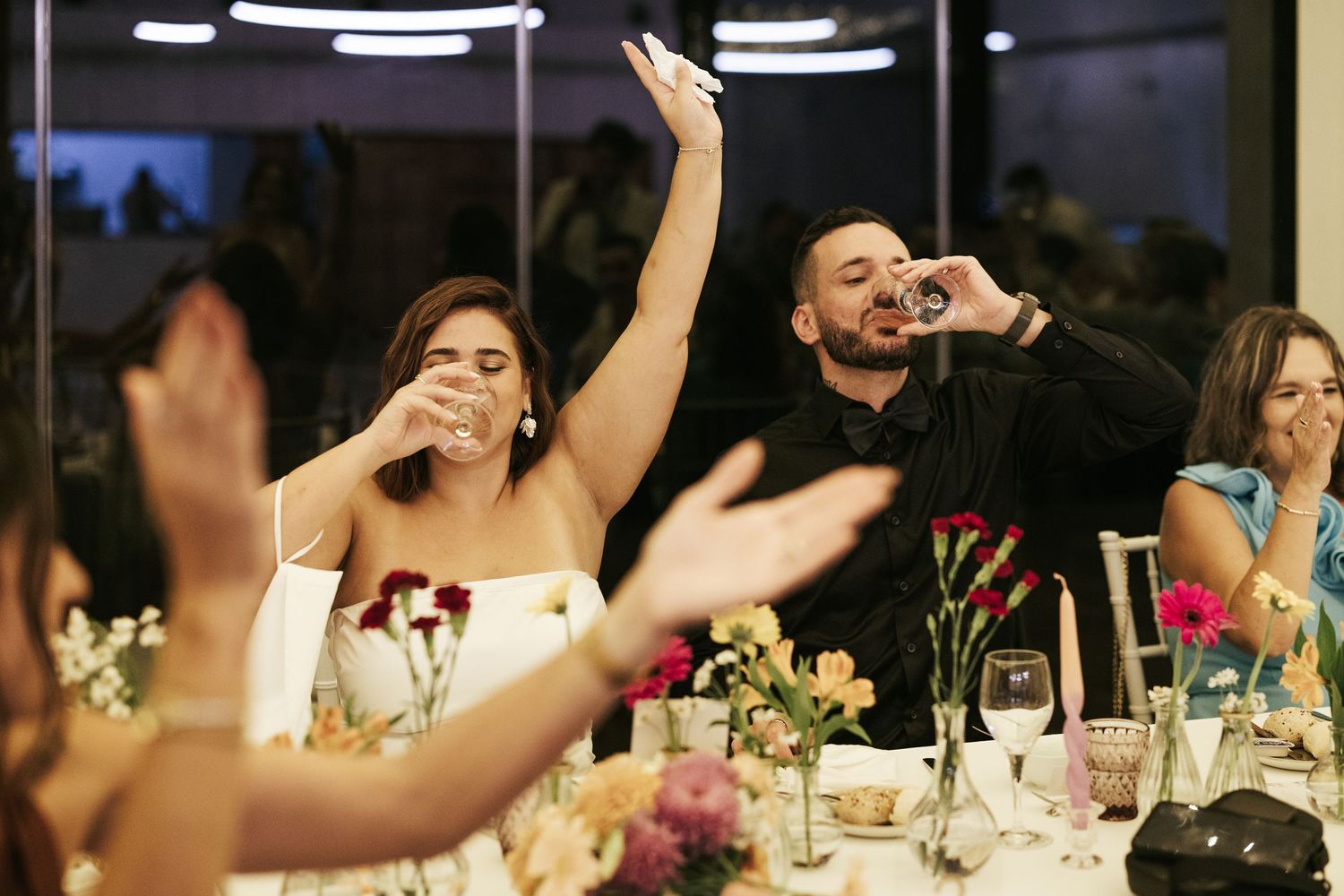Wedding guests raise their hands and cheer during an energetic reception dinner celebration at a darkened venue.