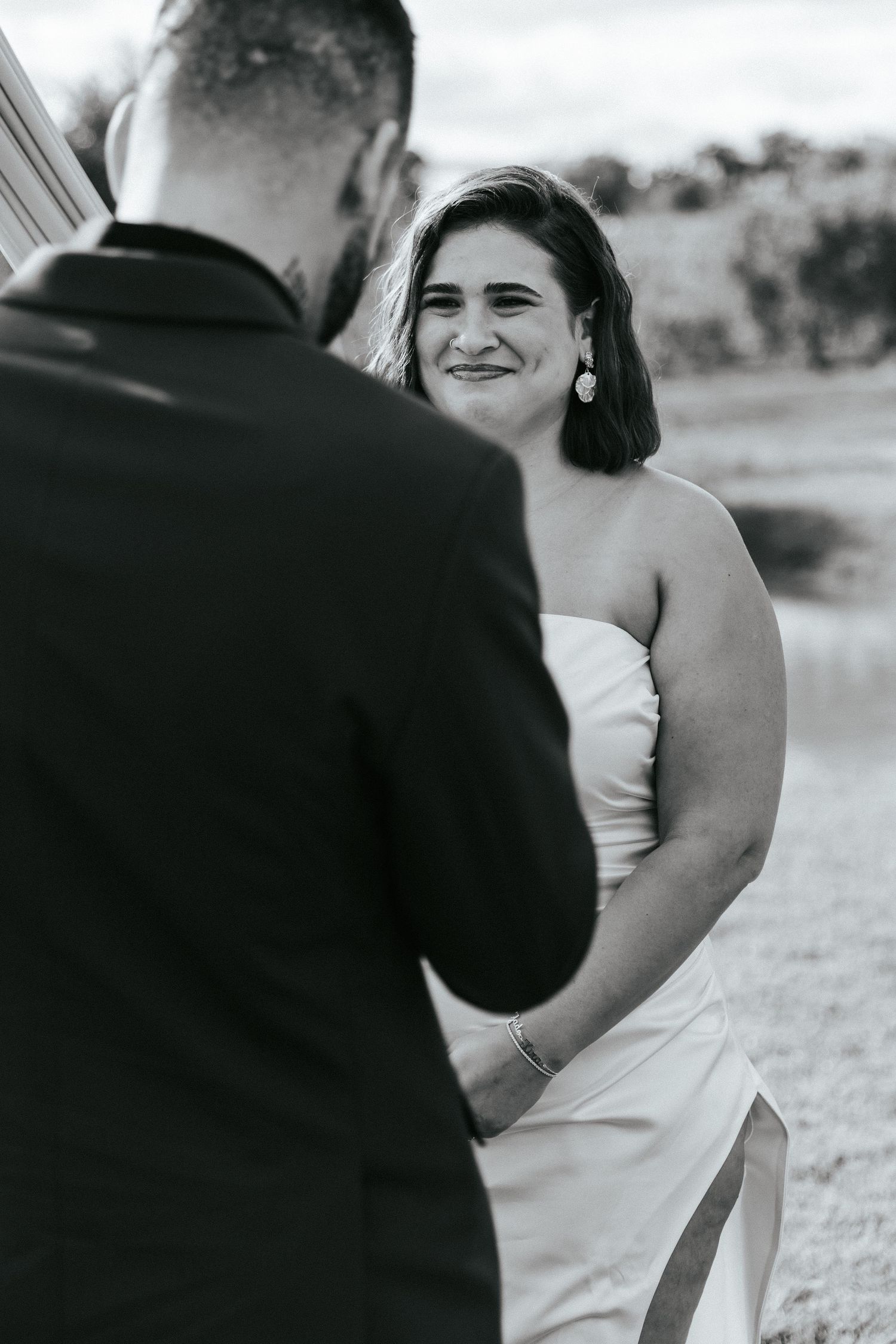 A black and white photo capturing a joyful moment between a couple wearing formal wedding attire outdoors.