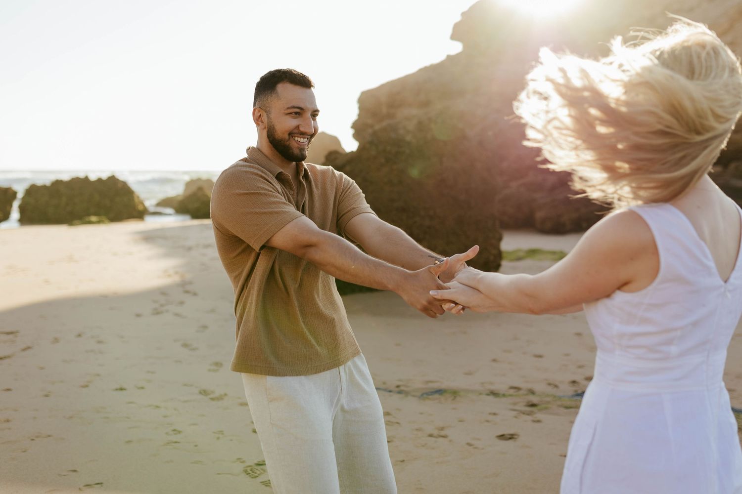 Two people dance together on a sandy beach at sunset with sunlight streaming behind them and ocean waves in the background.