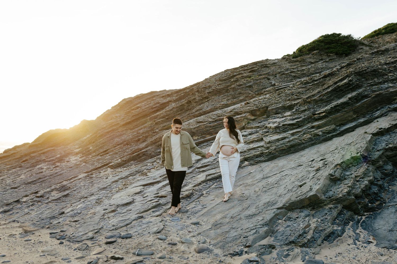 Two people walking together on rocky coastal terrain during golden hour sunset at the beach.