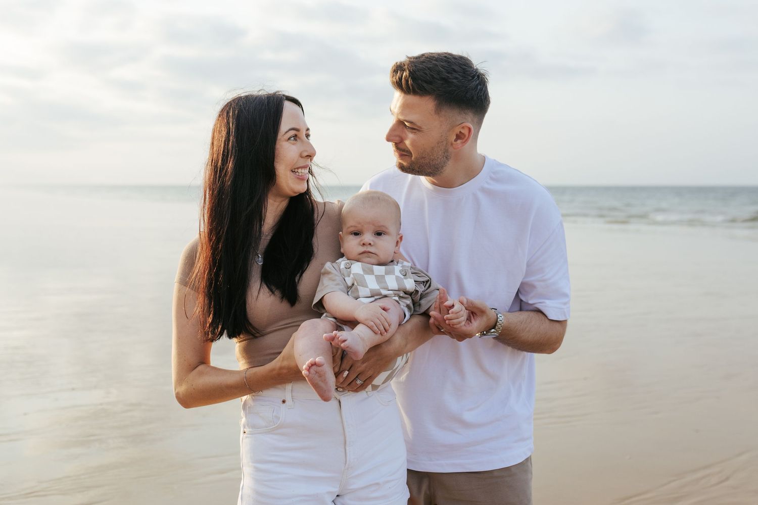 A couple wearing white outfits holds their baby while smiling on a sun-lit beach during sunset.