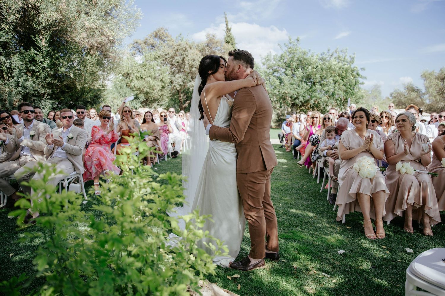 Couple shares their first kiss at outdoor garden wedding ceremony surrounded by guests on white chairs and greenery.