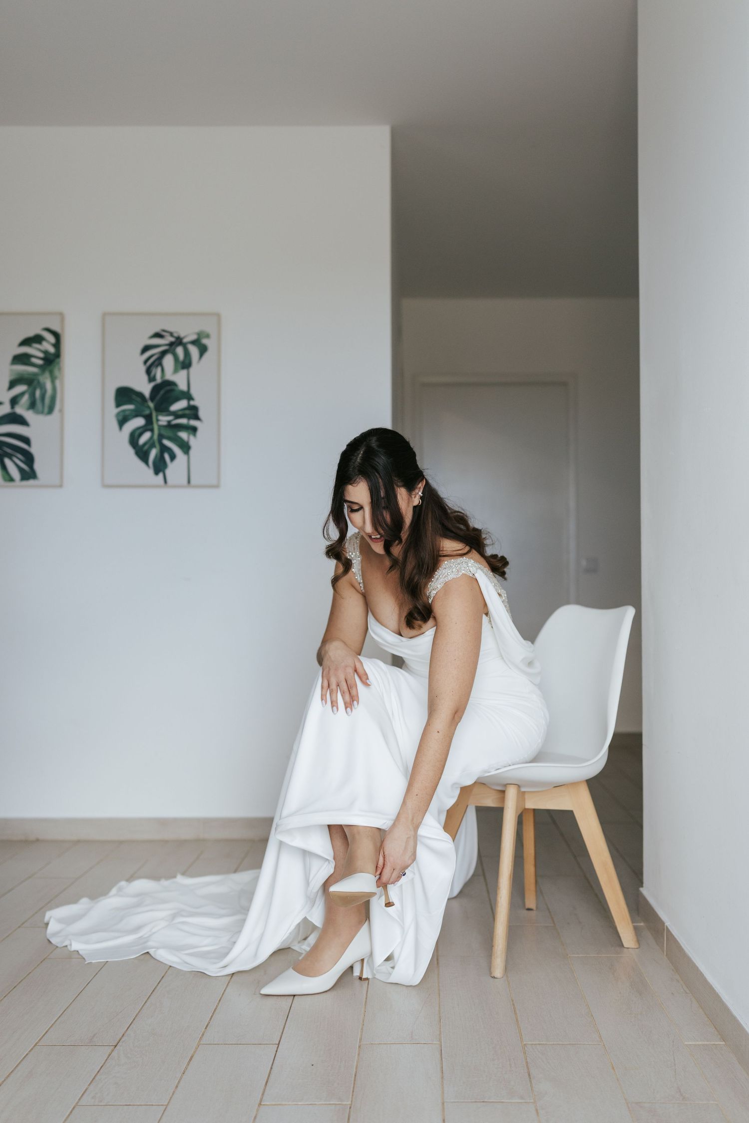 Elegant bride in white dress and heels sits on modern chair with botanical prints on wall in minimalist room.