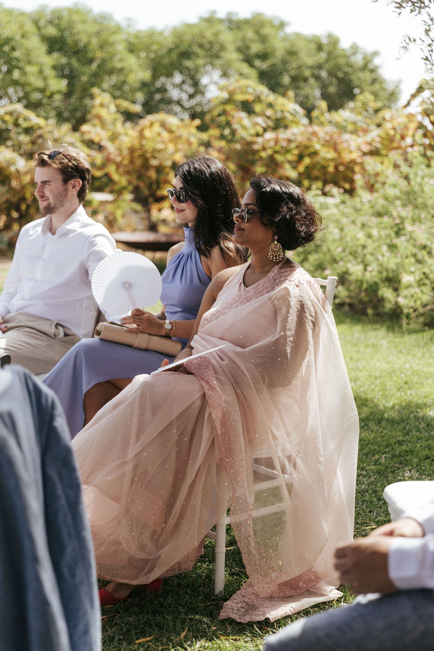 Guests seated outdoors at a garden wedding, woman in pink saree and sunglasses enjoying the ceremony.