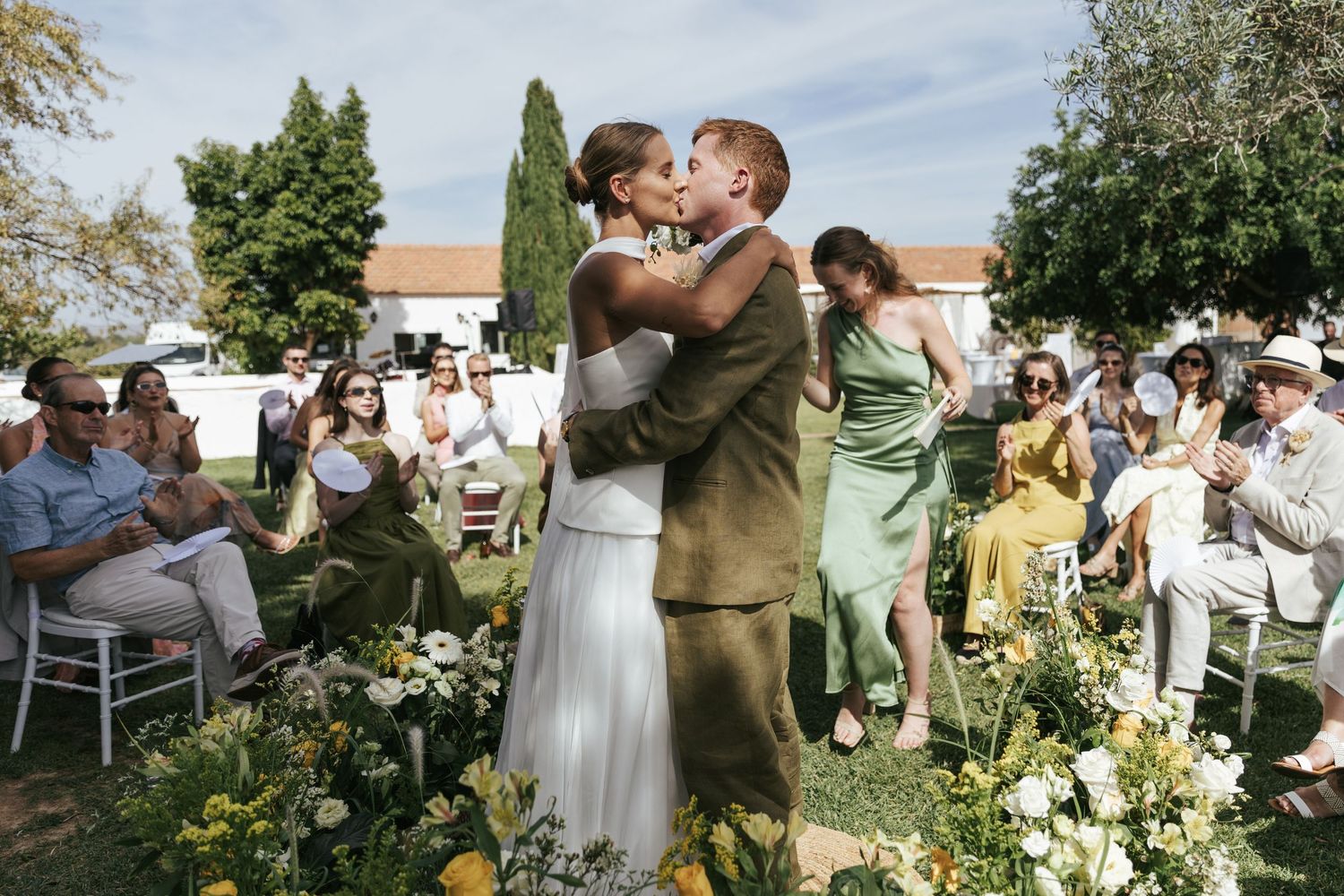 Bride and groom share a first kiss at an outdoor wedding ceremony surrounded by floral arrangements and seated guests.