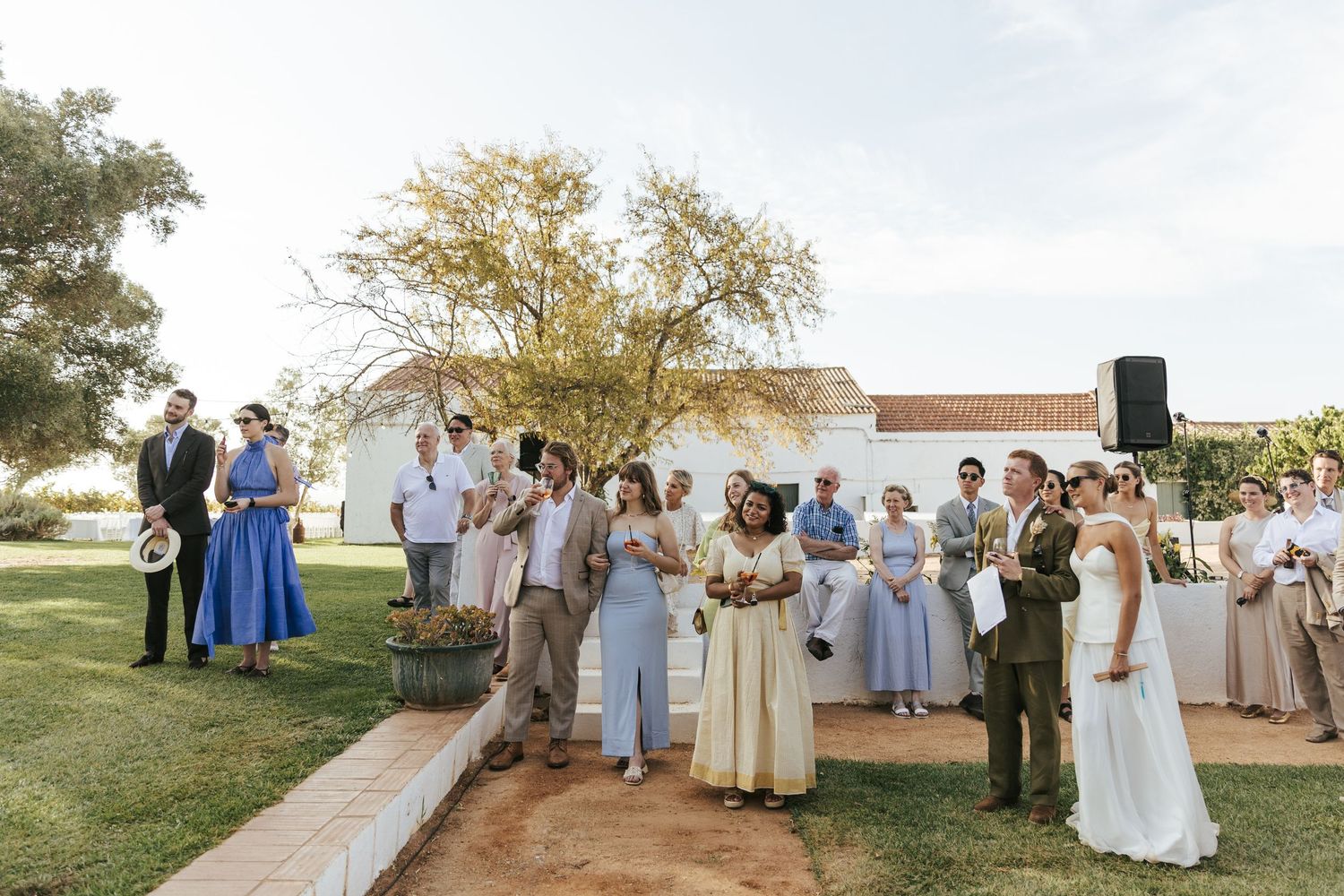 Wedding guests gathered outdoors on green lawn near white chapel, celebrating in elegant attire on a sunny day.