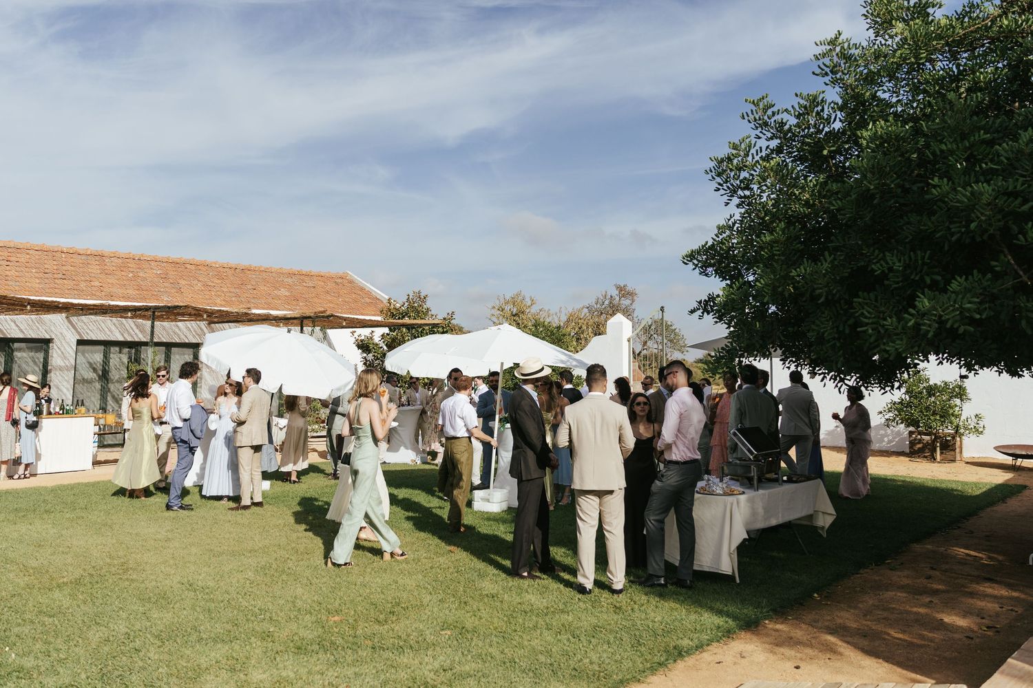 Outdoor wedding cocktail hour with guests mingling under white umbrellas on a sunny day.