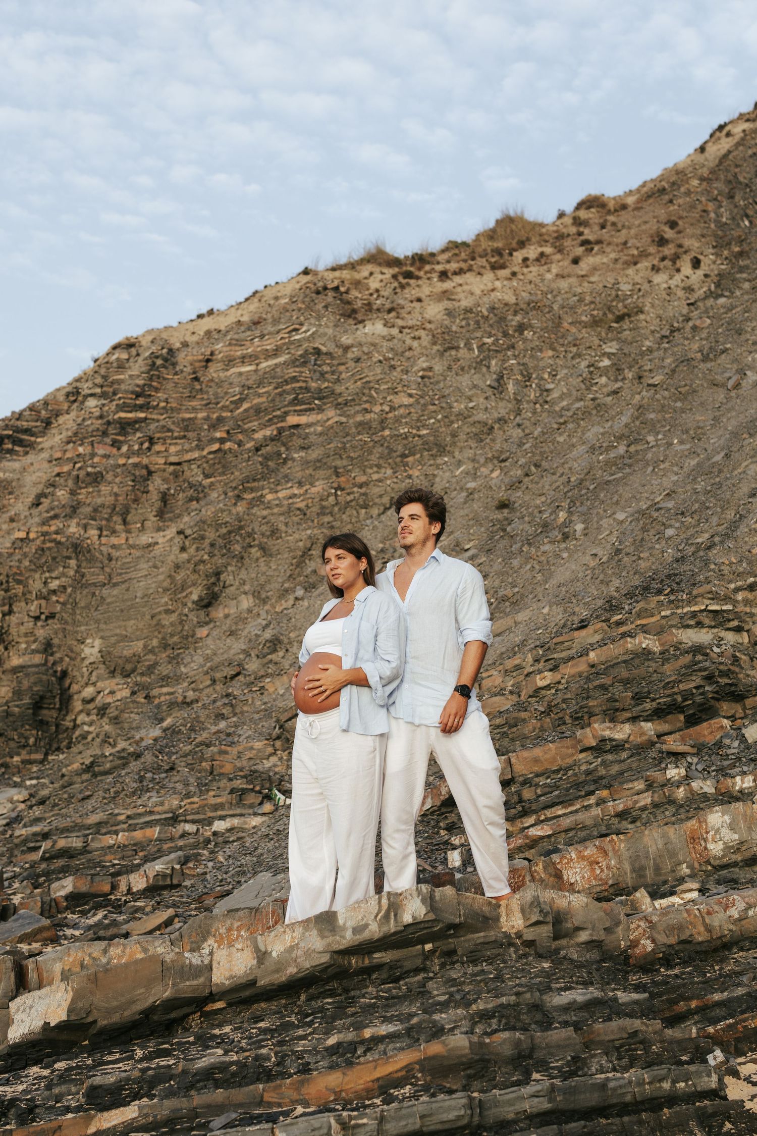 People in white outfits stand against a dramatic rocky cliff face at sunset creating a striking silhouette.