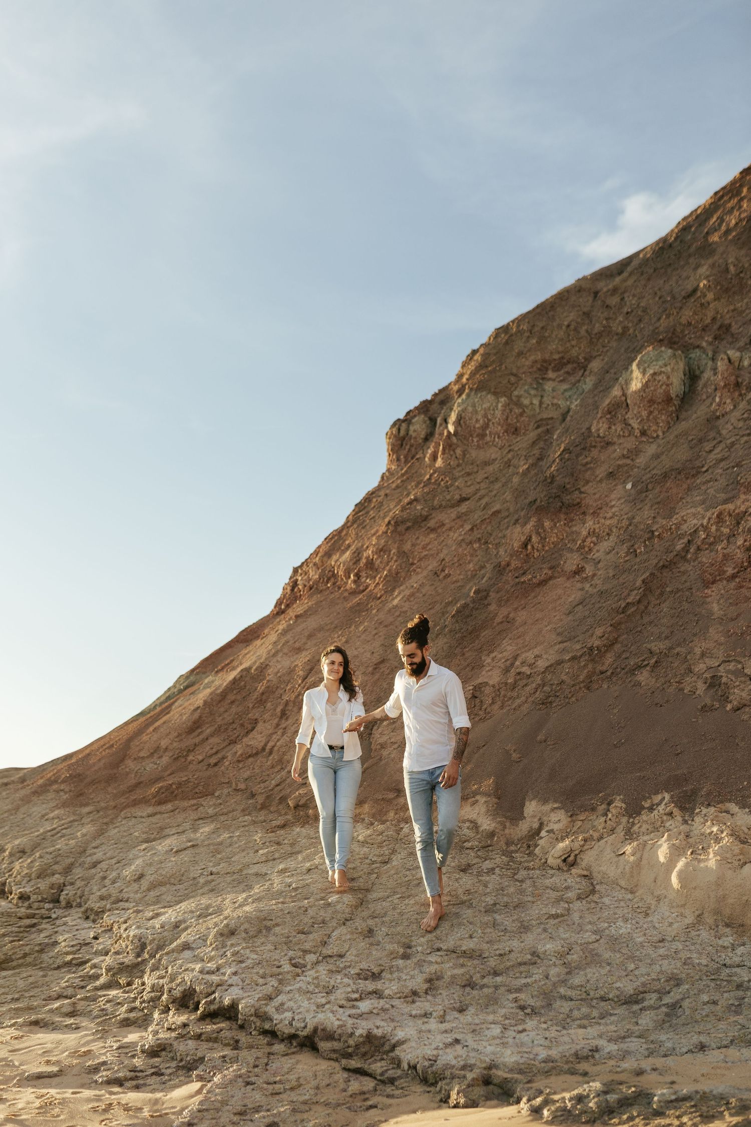 Two people in casual white outfits walk together along a rugged coastal cliff during golden hour.