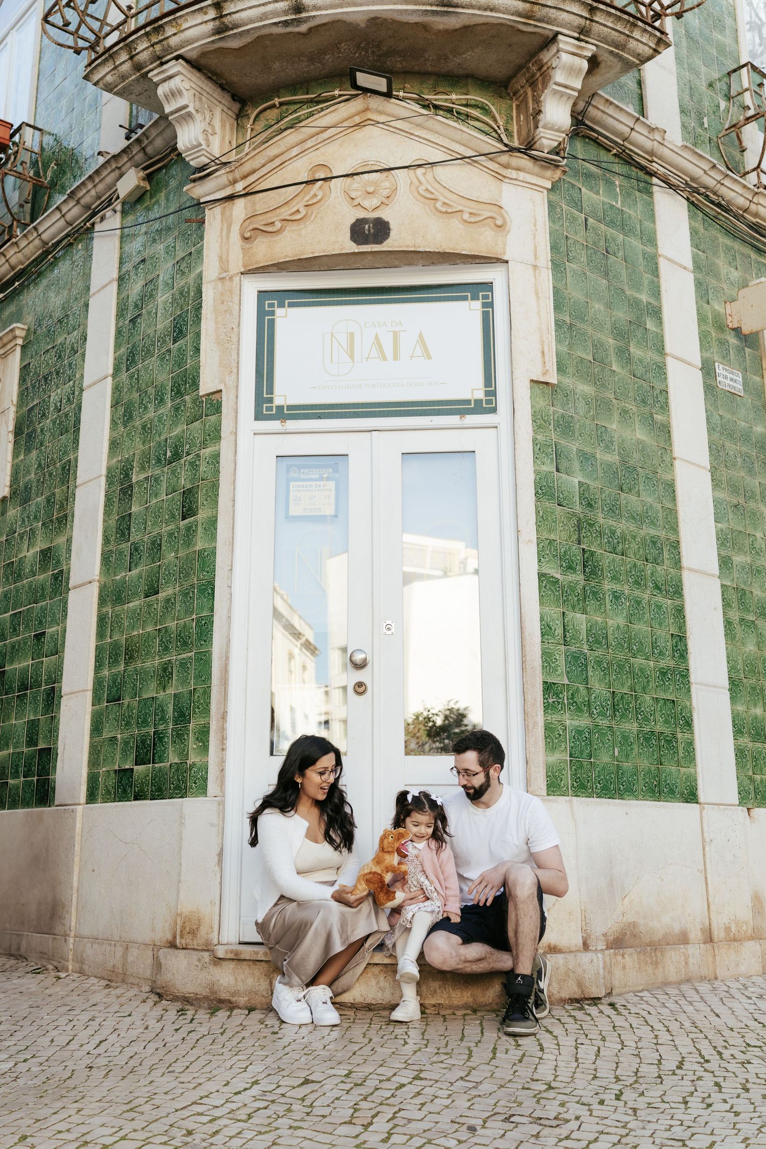 A family sits with their small dog in front of a vintage building with green tiled walls and ornate white doorway.